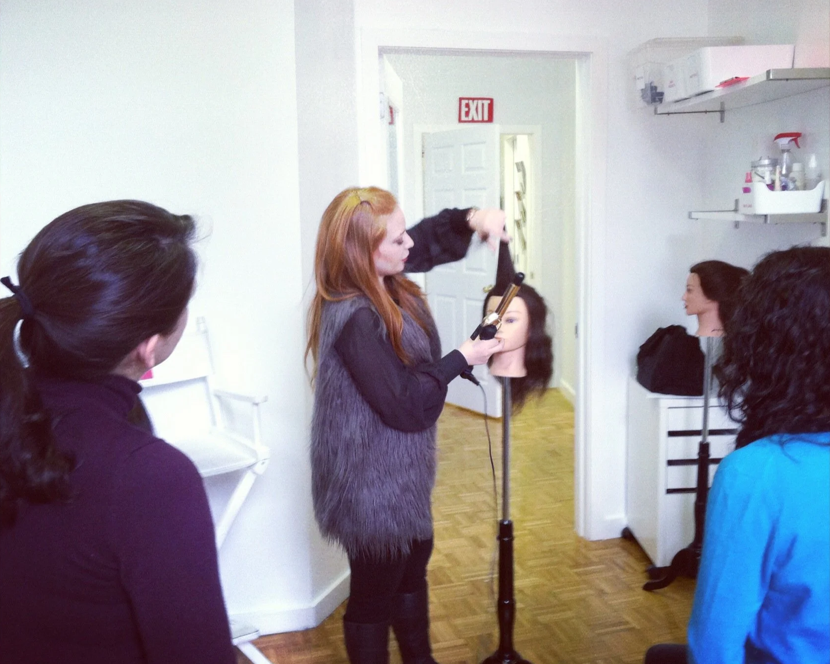 A hairstyling class with a woman teaching students how to style wigs, using a mannequin head on a stand, in a room with white walls and shelves.