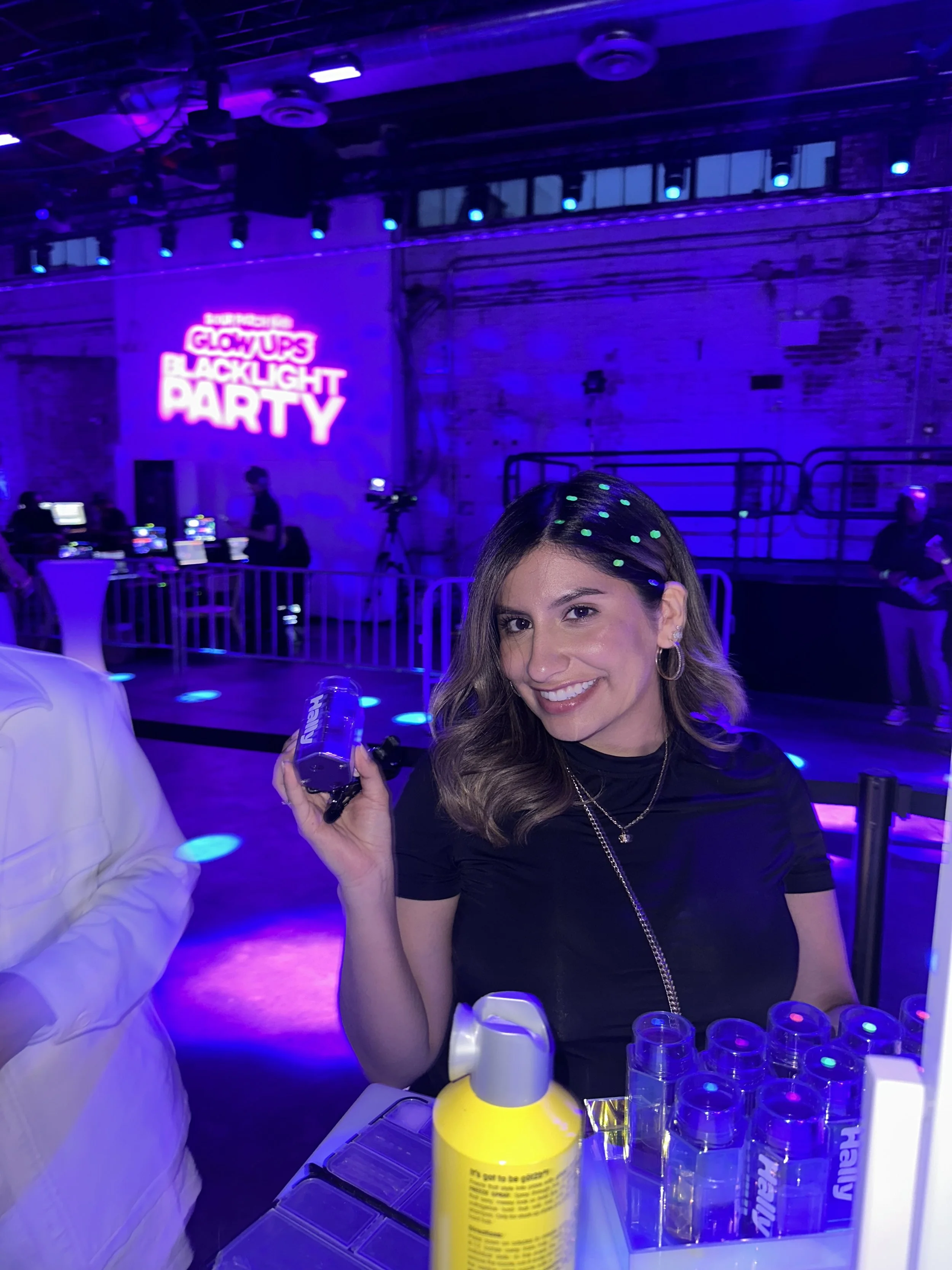 A young woman with shoulder-length hair smiling and holding a small purple item, sitting at a table with bottles of spray on it. She has bright green dots in her hair and is wearing hoops, a black turtleneck, and layered necklaces. The background features a neon sign that reads 'Glow Up's Blacklight Party' and is decorated with purple and blue lighting, with other people and equipment visible.