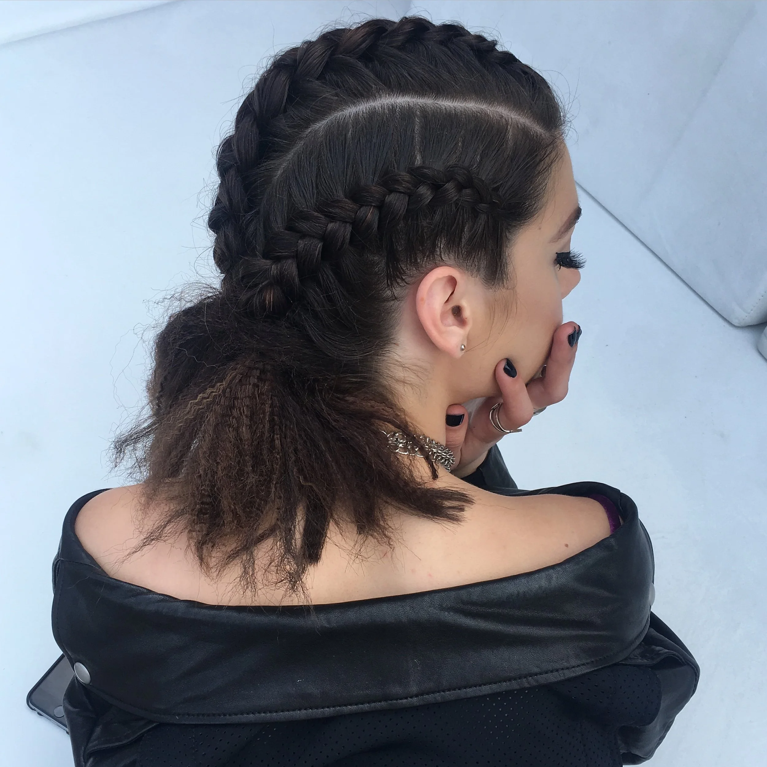 A woman with dark brown hair styled in two French braids, with the rest of her hair curly and loose, sitting with her hand covering her mouth. She is wearing a black leather jacket and has multiple rings and earrings.