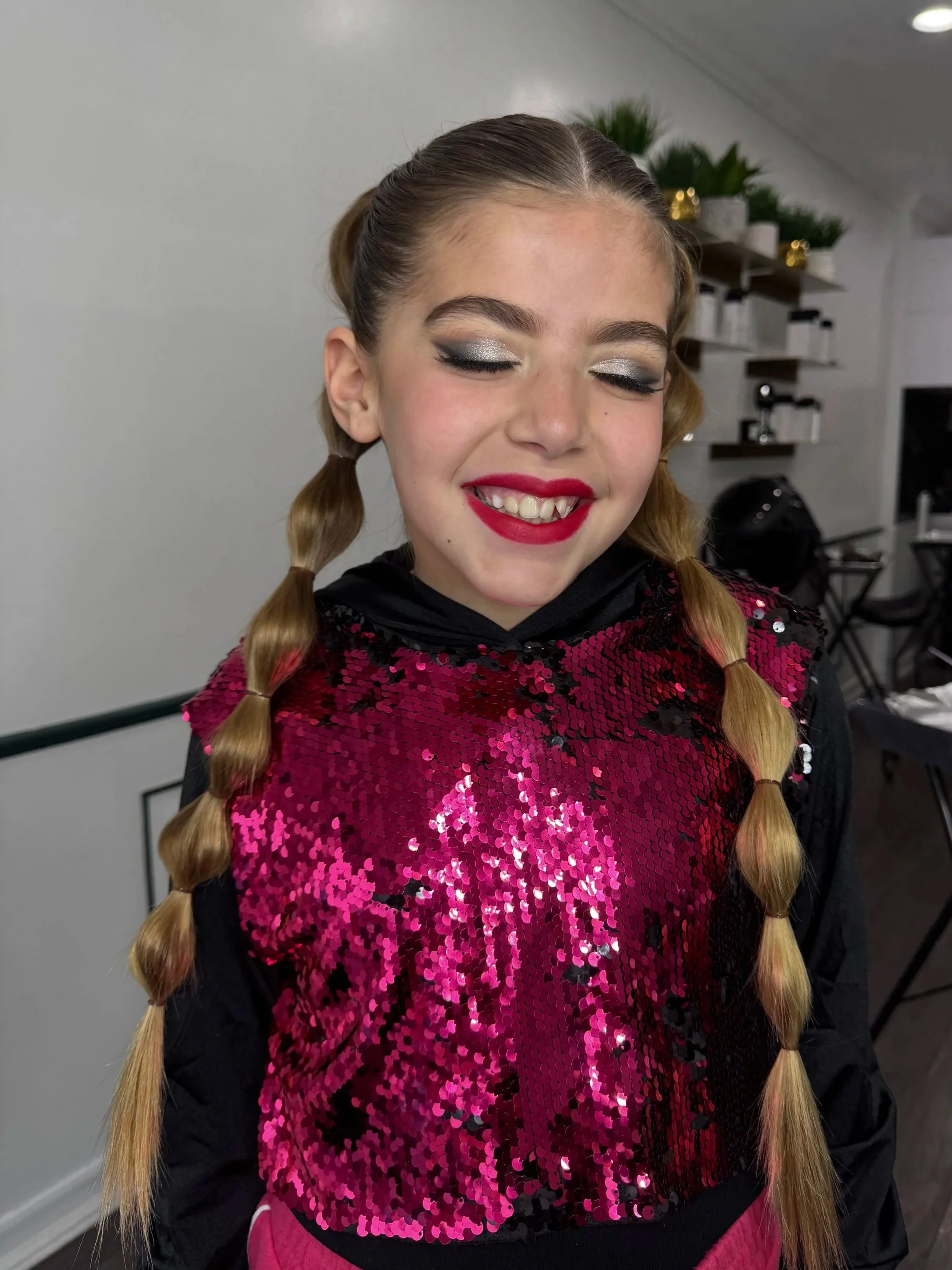 Smiling girl with double braided hair, wearing silver eye makeup and red lipstick, dressed in a black hoodie with pink sequined design on the front, in a room with shelves and potted plants in the background.