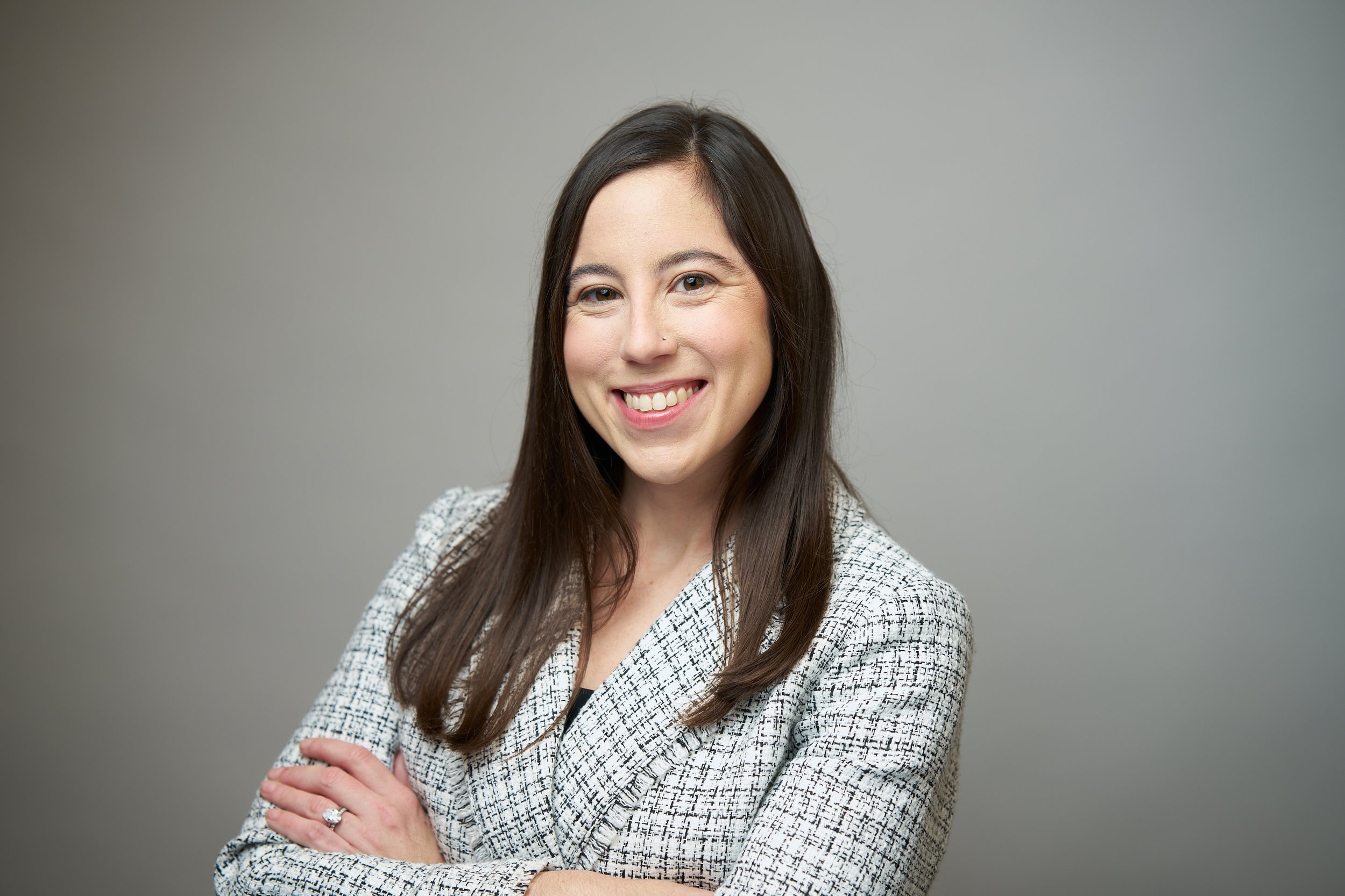 A woman with long dark brown hair, smiling, wearing a black top and a checked blazer, standing against a plain gray background.