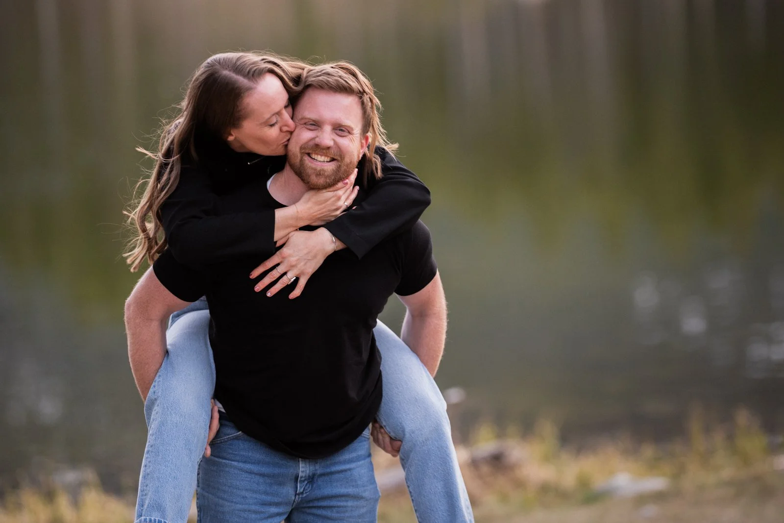 Playful couple sharing a piggyback ride during a lakeside engagement session in Red Lodge Montana