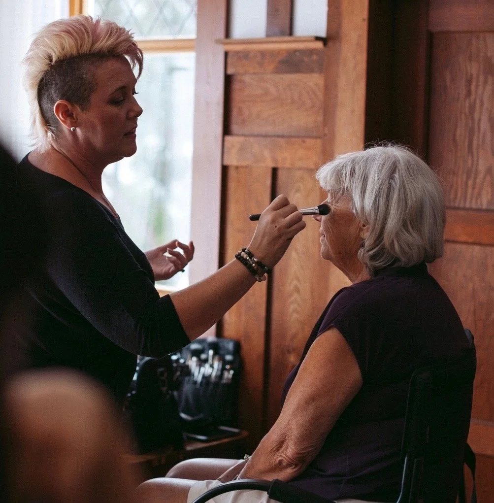 Makeup artist applying foundation to a mature client's face near a window