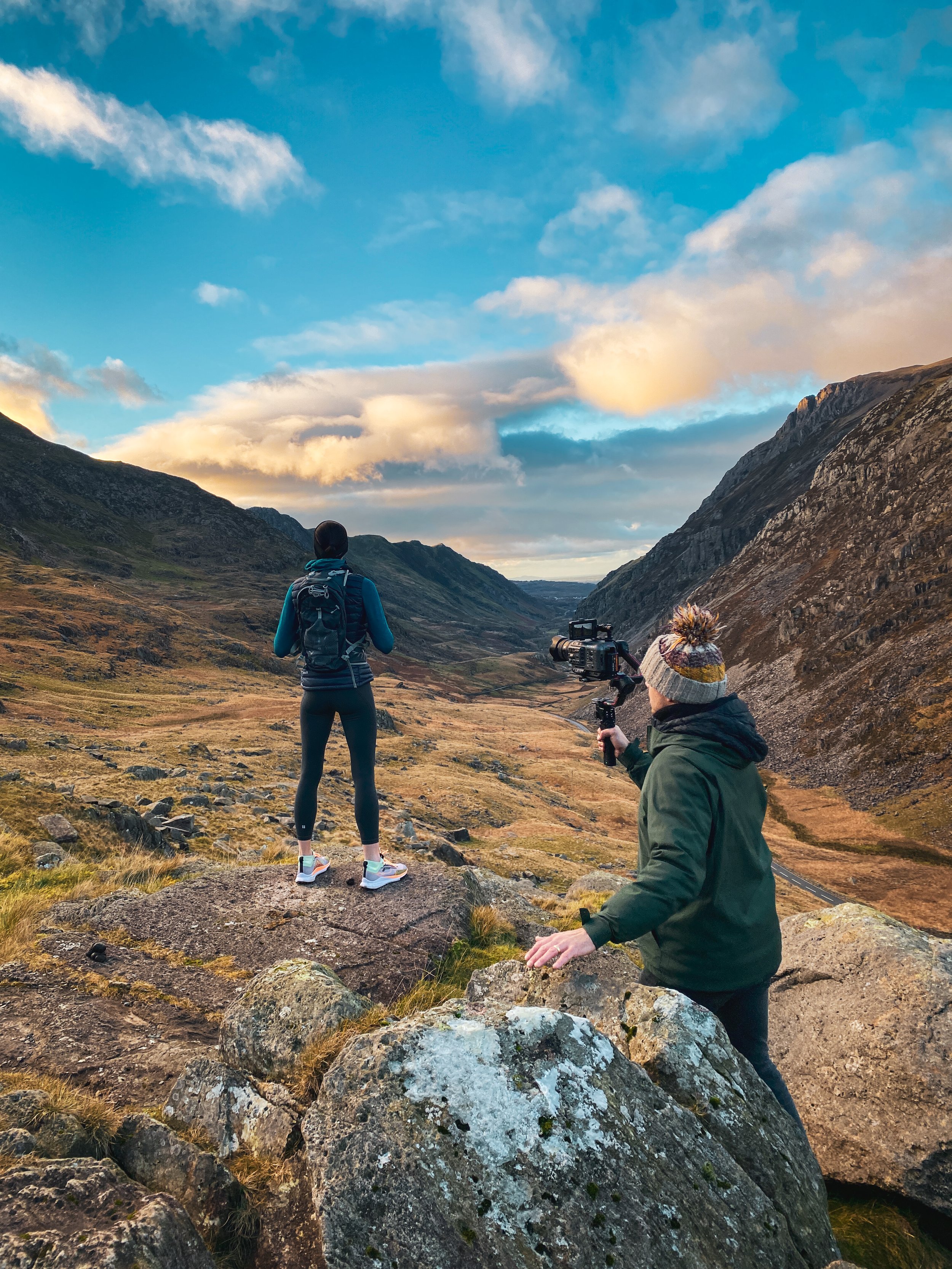 Sri Southall DOP filming a model with a Ronin RS3 gimbal and Sony FX3 camera while she stands on rocks, overlooking a scenic mountain valley during sunset or sunrise.