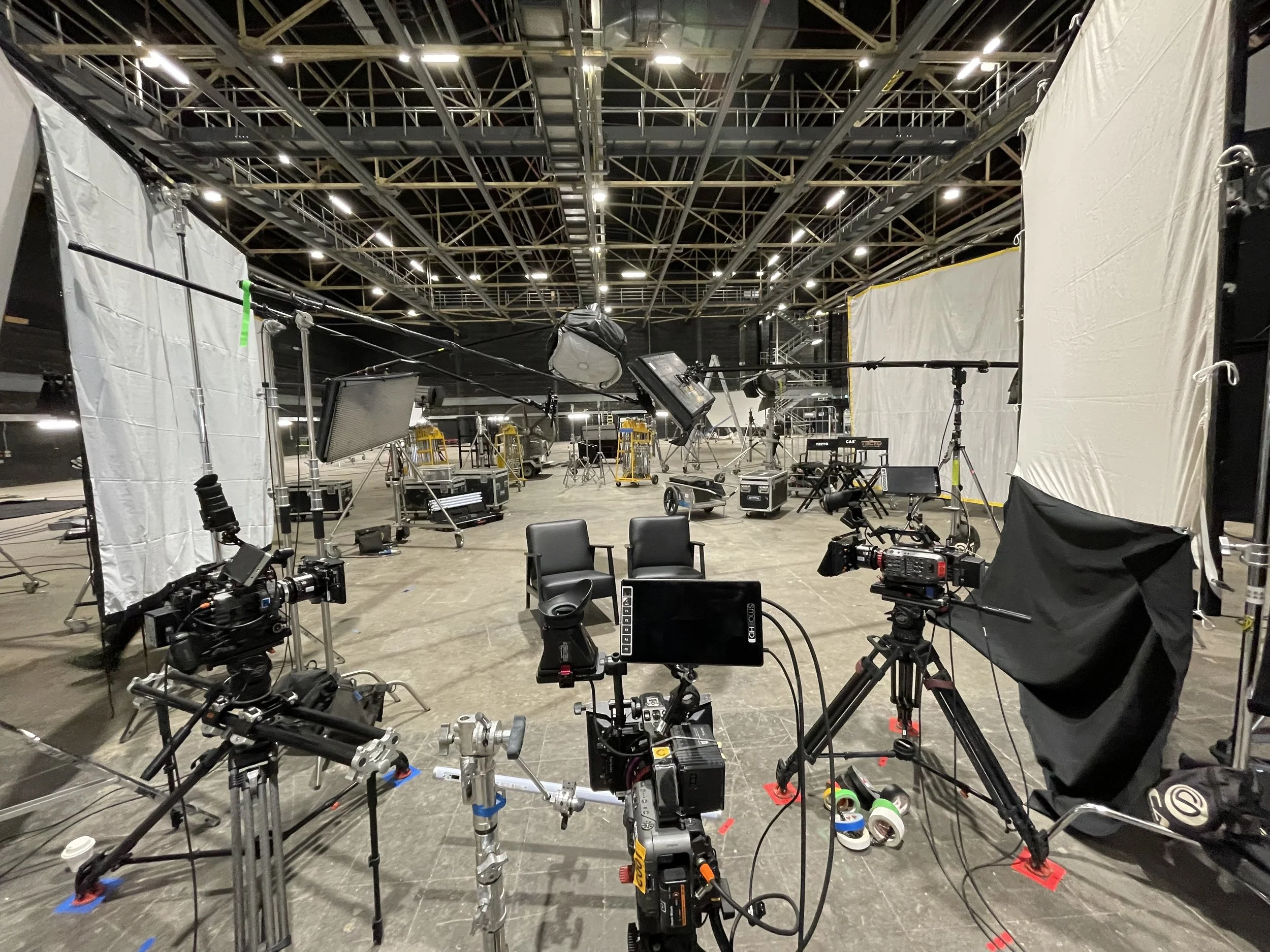 An empty television or film studio set with cameras, lighting equipment, chairs, and backdrop curtains, ready for filming or a photography shoot.