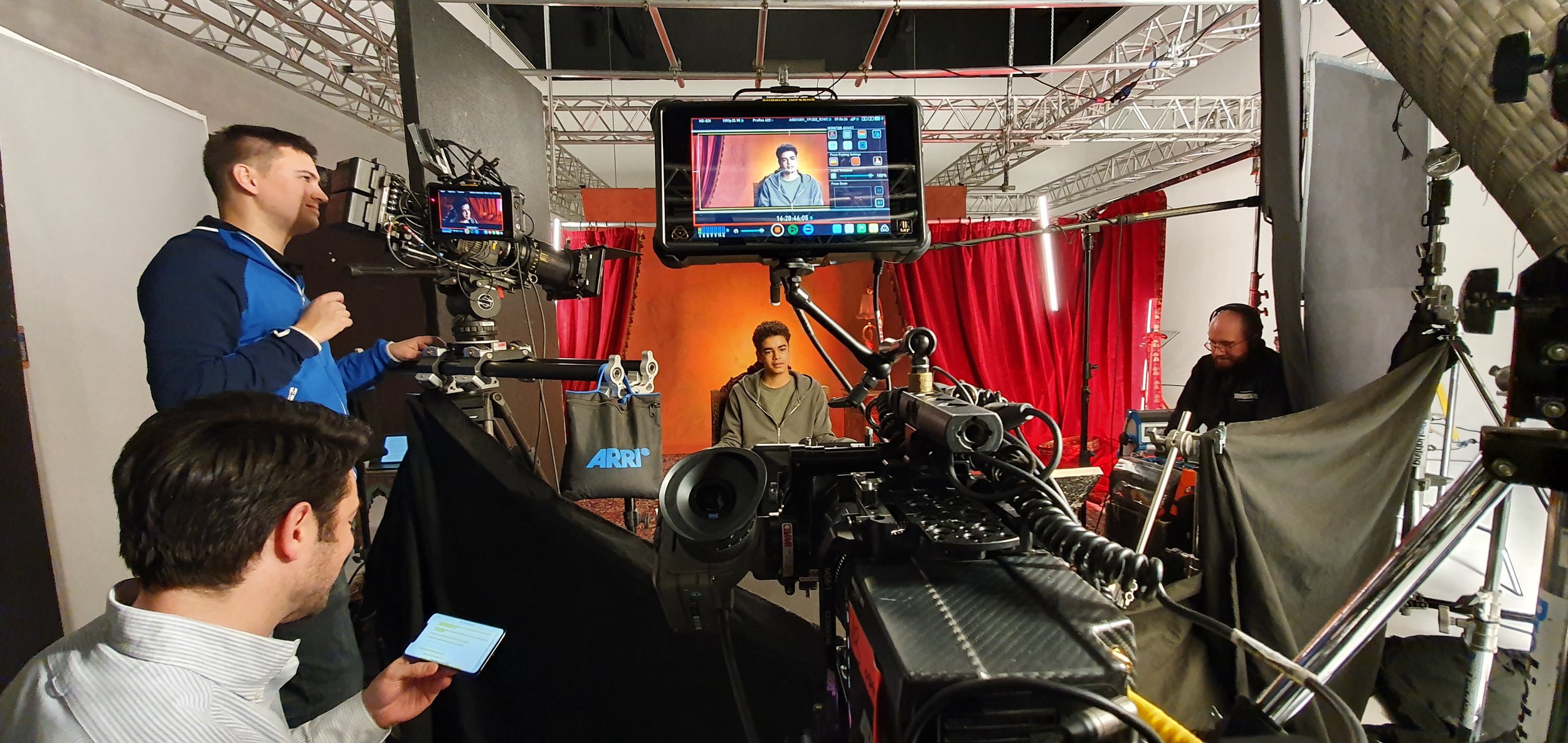 Inside a film or video production set, a young man is seated in front of a red curtain backdrop, being filmed. Crew members and equipment, including cameras, monitors, and lighting, surround him, with one crew member operating a camera on the left and another on the right adjusting lighting. A crew member in the foreground is looking at a phone, and a console with controls is visible in the foreground.