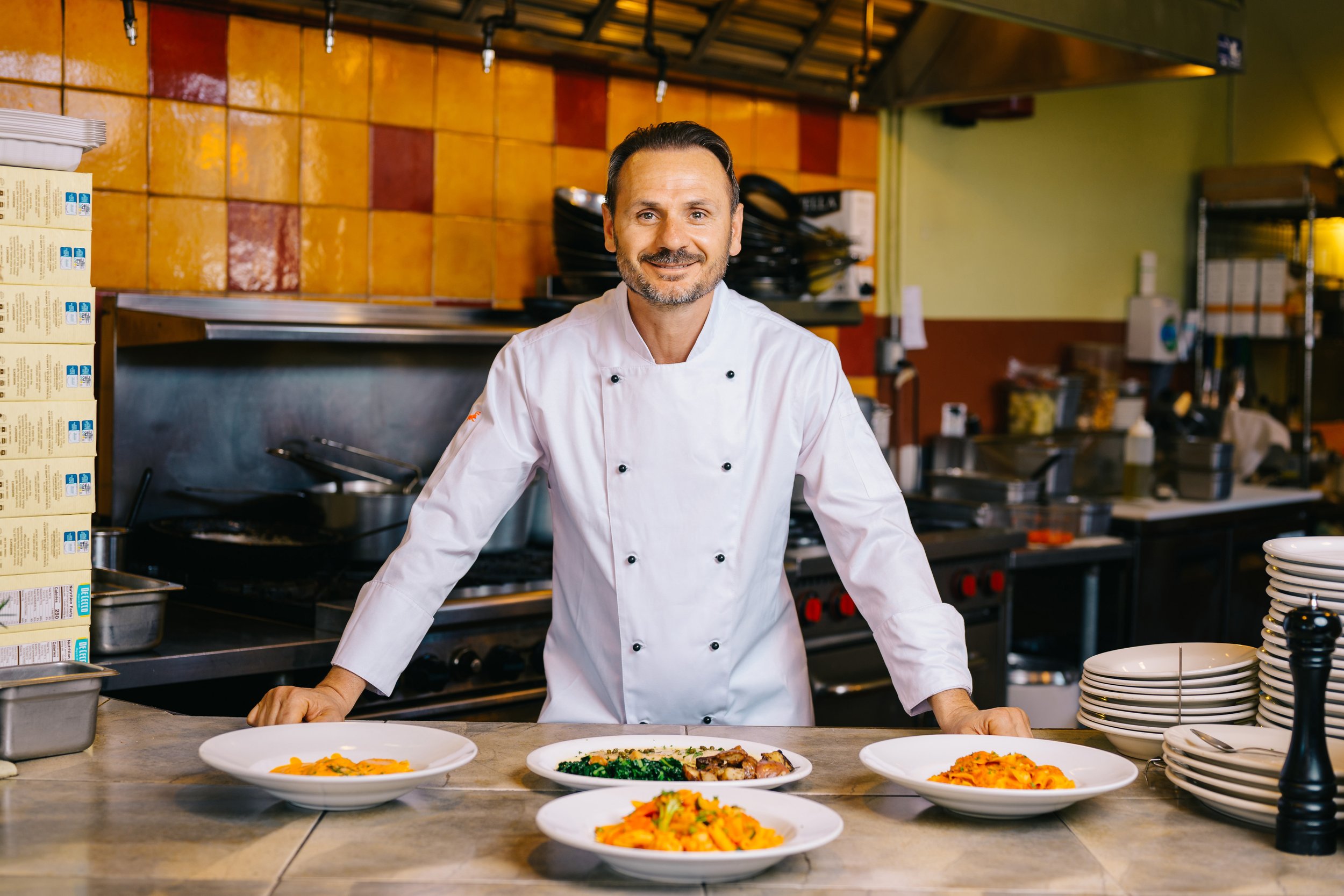 Chef Luca standing in his restaurant kitchen behind four dishes.
