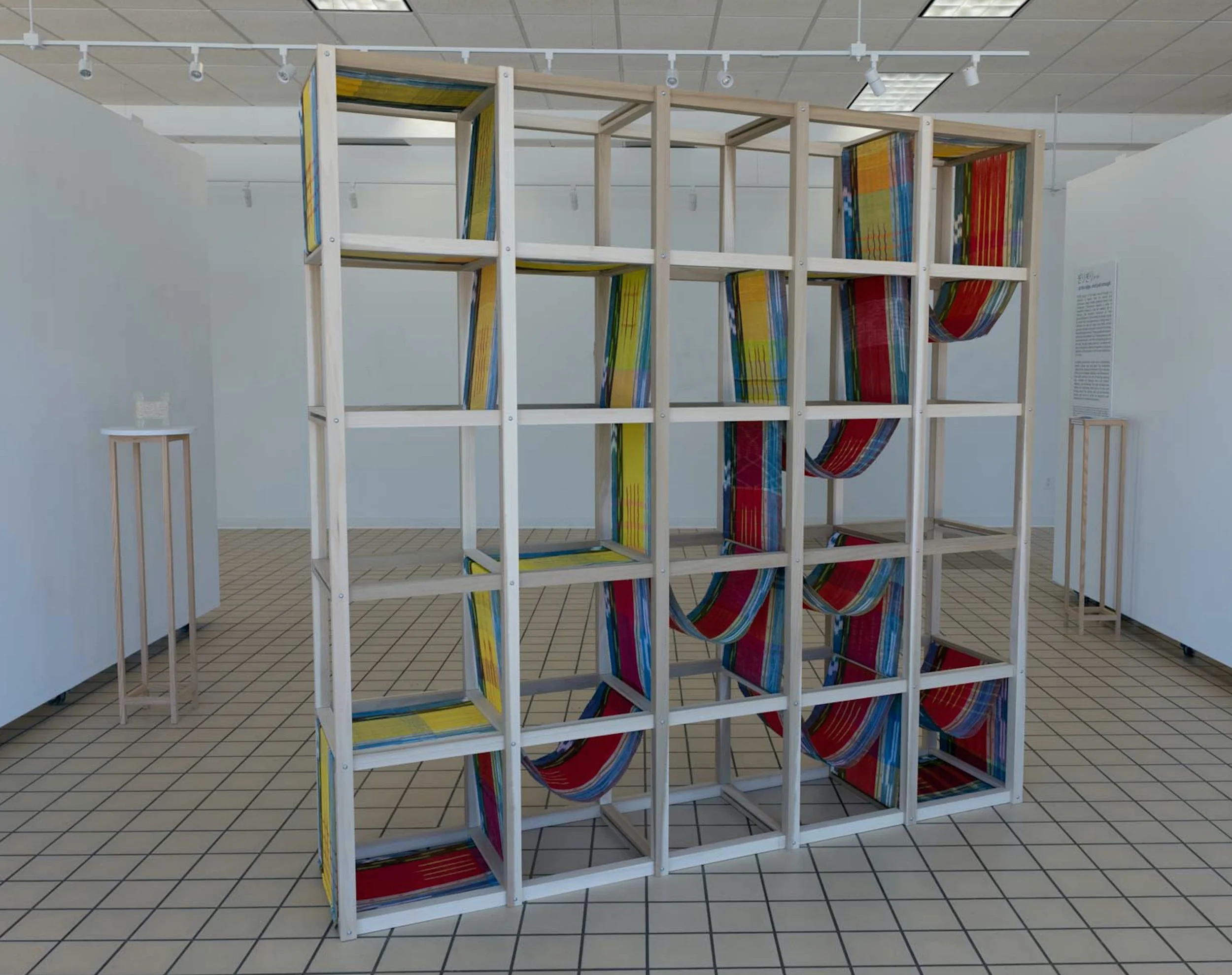 View of exhibition space. At the left, a pedestal holds a small cast glass sculpture. At the center, a large 7'x7'x18" sculpture fills most of the space. The wood frame holds 60' of handwoven kasuri dyed fabric. The long warp contains 4 stripes.