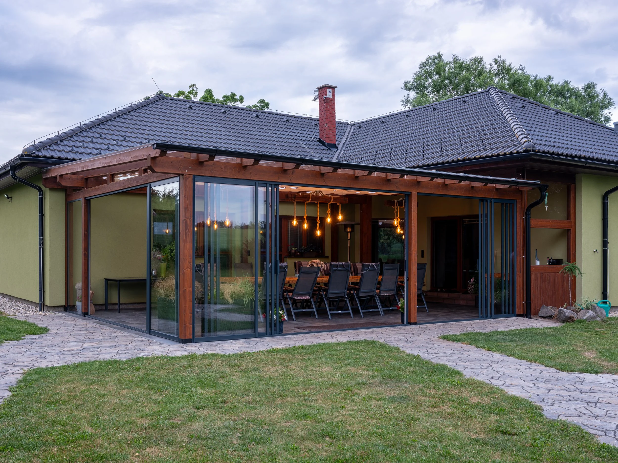 A house with a green exterior, black tiled roof, and an enclosed glass patio area with a long dining table and chairs inside. The patio features wooden beams and hanging light bulbs. There is a stone pathway leading to the house and a well-maintained