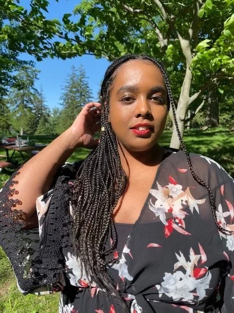 Norah is standing in a park with the sky, trees and a picnic table behind them.
