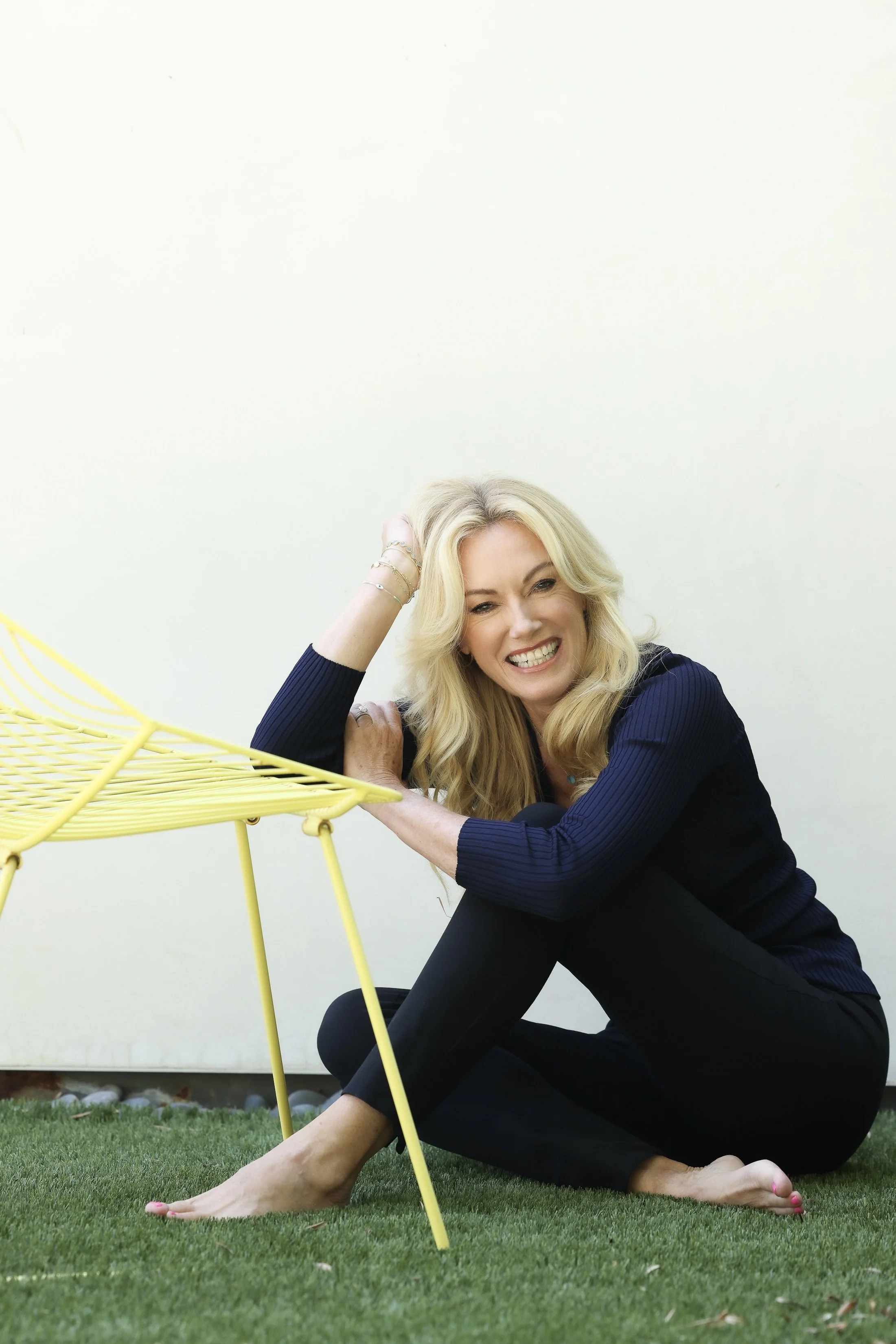 A woman with long blonde hair sitting on the grass, smiling at the camera, with yellow outdoor furniture nearby, against a plain white background.