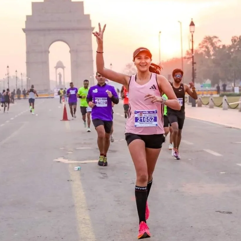 A woman running in a marathon near the India Gate, smiling and making a peace sign, with other runners behind her and the sunrise in the background.