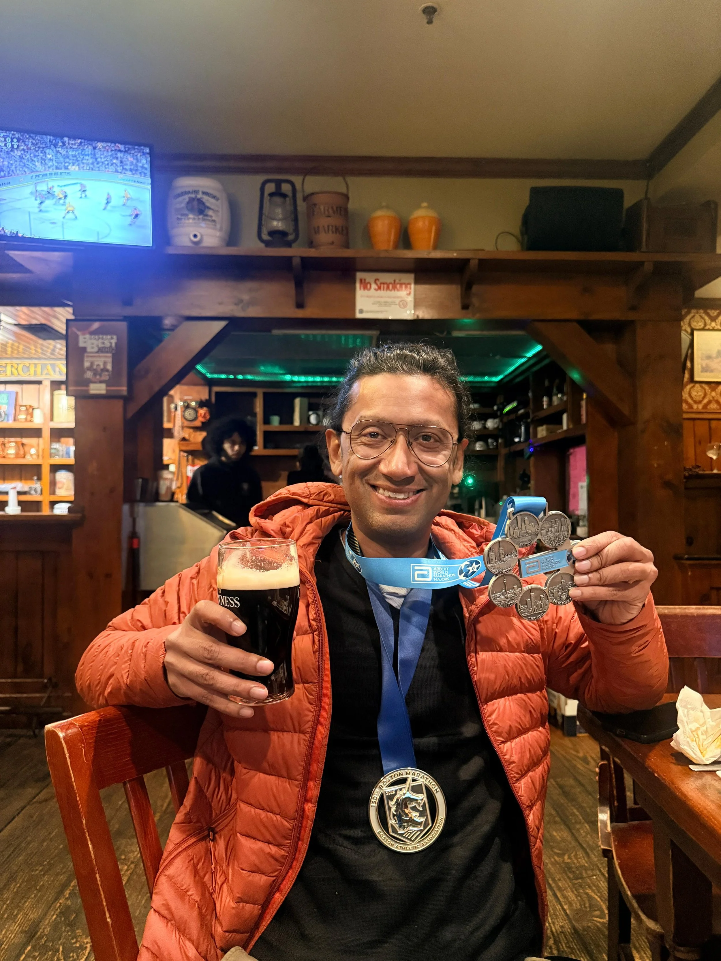 A man wearing glasses and a black shirt with a Boston Marathon medal around his neck, holding a glass of dark beer and showing several medals with a medal ribbon, sitting in a cozy restaurant or pub with wooden decor.
