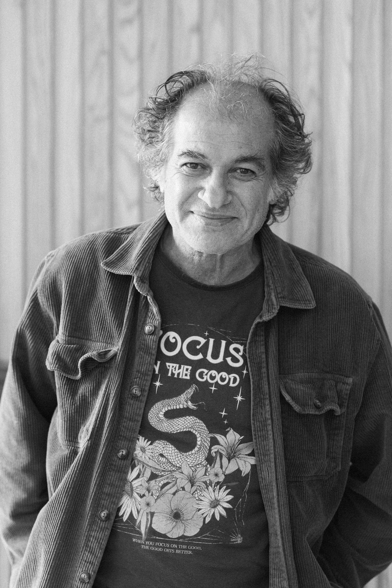 Black and white photograph of a man with curly hair wearing a graphic t-shirt with a snake and flowers design, and an unbuttoned corduroy jacket, standing in front of a wooden panel background.