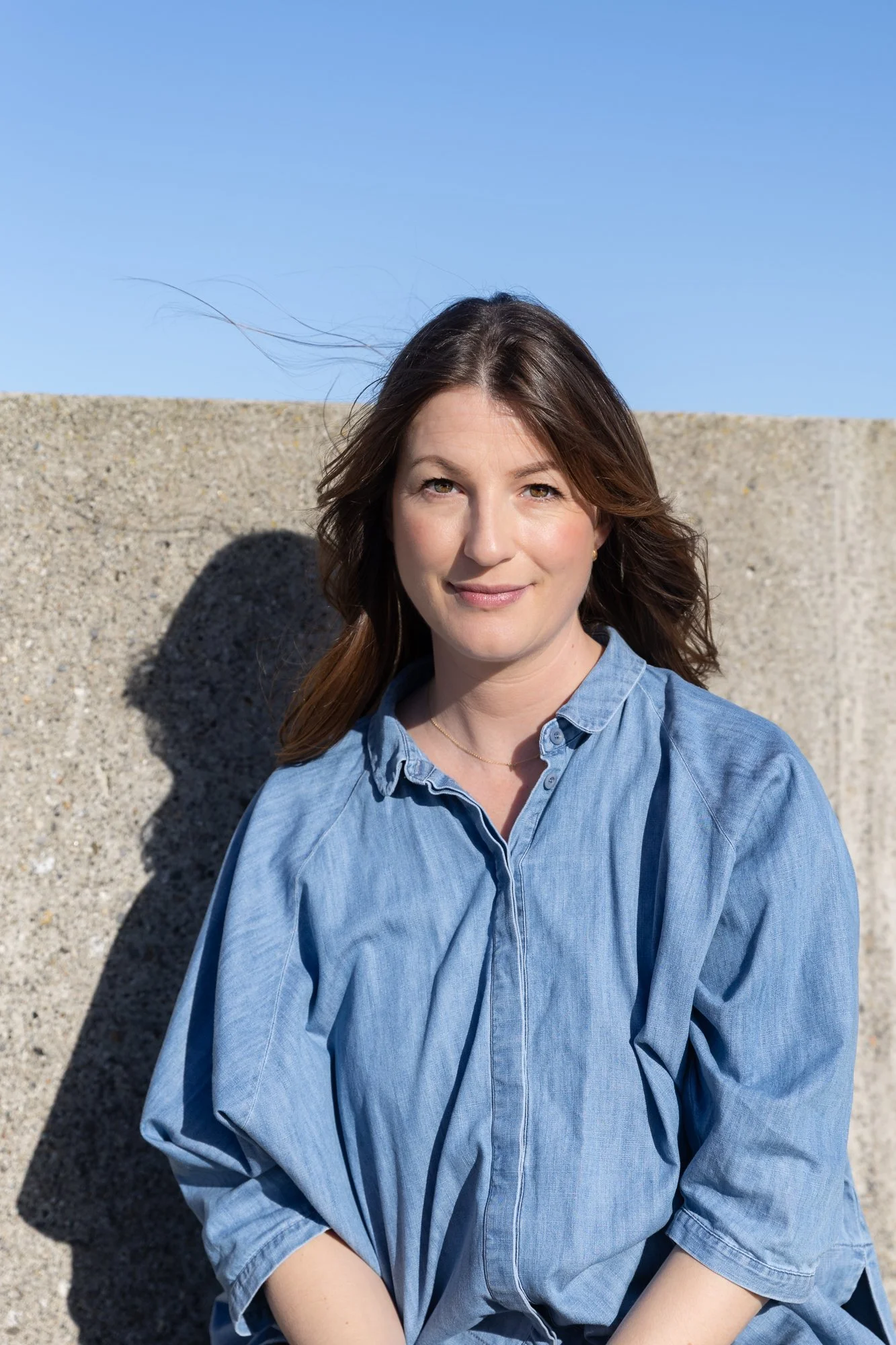 A woman with brown hair wearing a blue denim shirt standing outdoors against a concrete wall and blue sky, with sunlight casting shadows.
