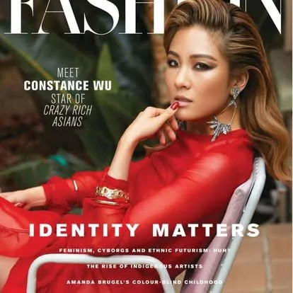 Magazine cover featuring a woman with long, wavy hair wearing a red outfit with jewelry, sitting outdoors among green plants, with text about fashion and identity.