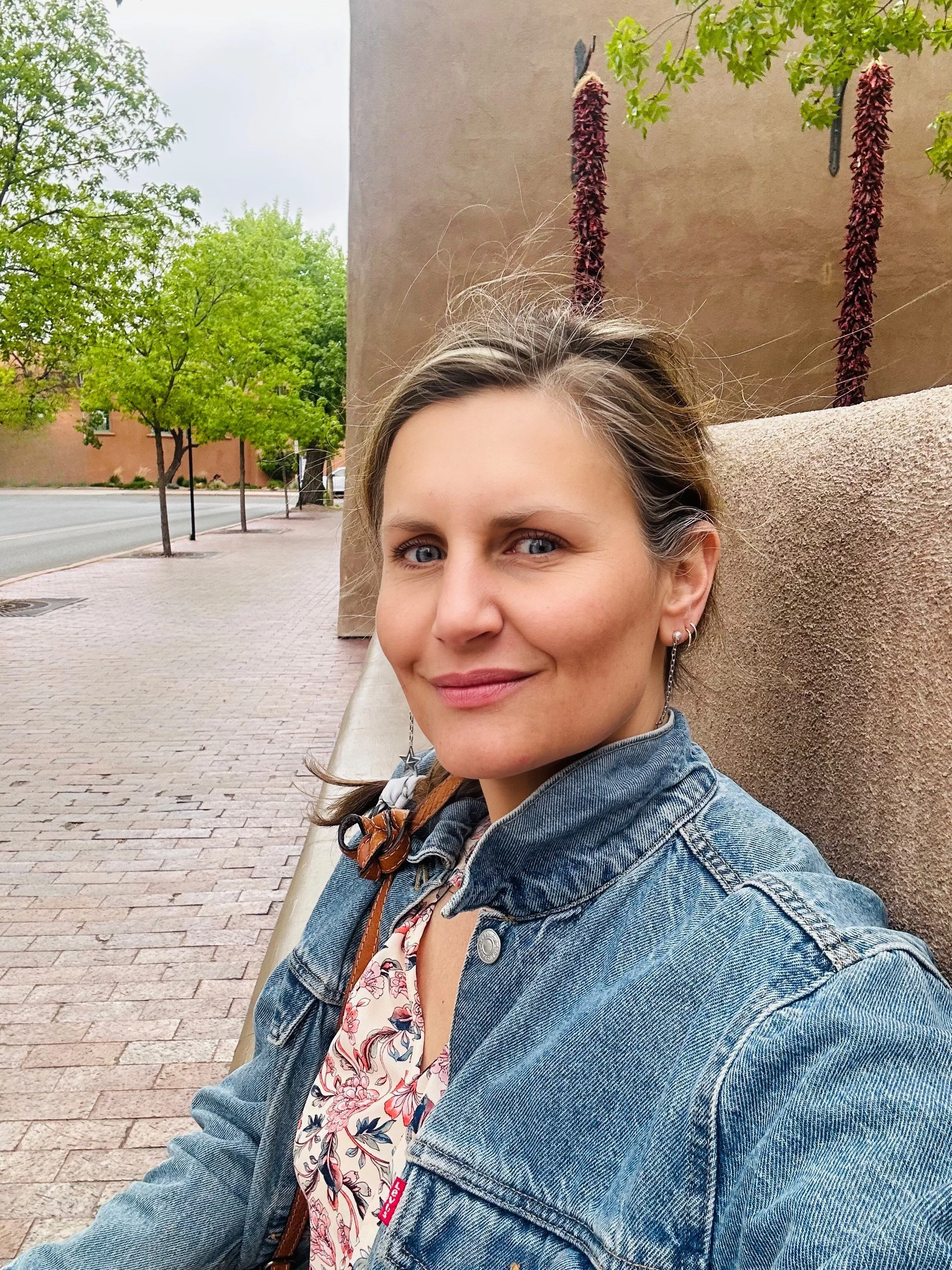 A woman sitting on a bench in an outdoor urban setting with trees, brick sidewalk, and a building wall behind her. She has light brown hair, blue eyes, and is wearing a denim jacket, earrings, and a floral top.