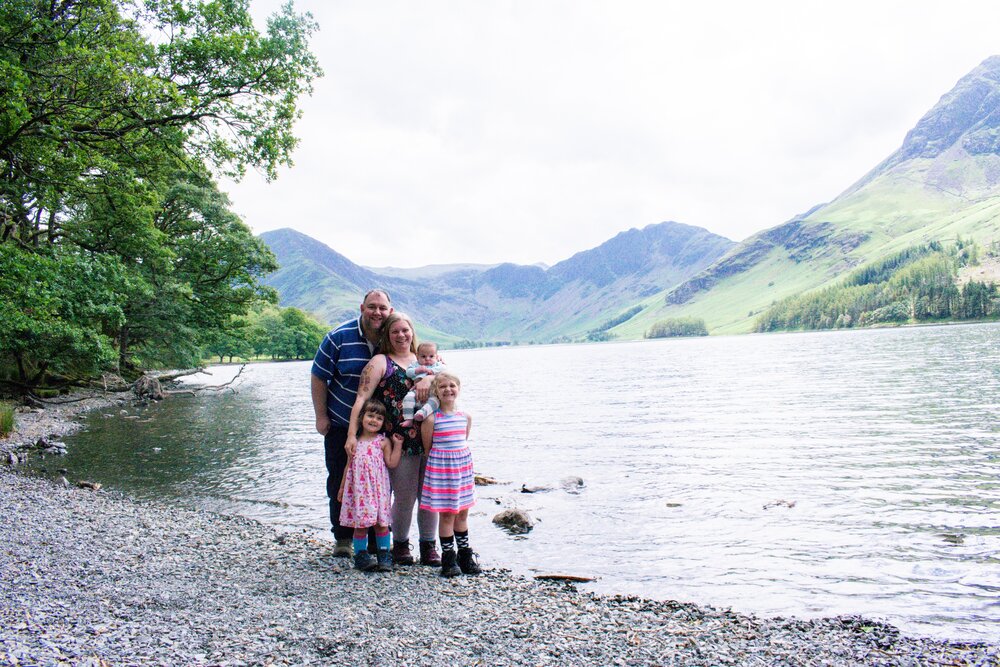 The Hassan family are stood together on the lakeshore of Buttermere with the lake and lakeland fells behind them.