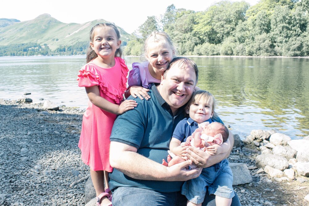 Pickle and Squidgy are stood on a rock behind John who has Munchkin on his lap with Peanut. They are on the lakeshore of Derwenwater.