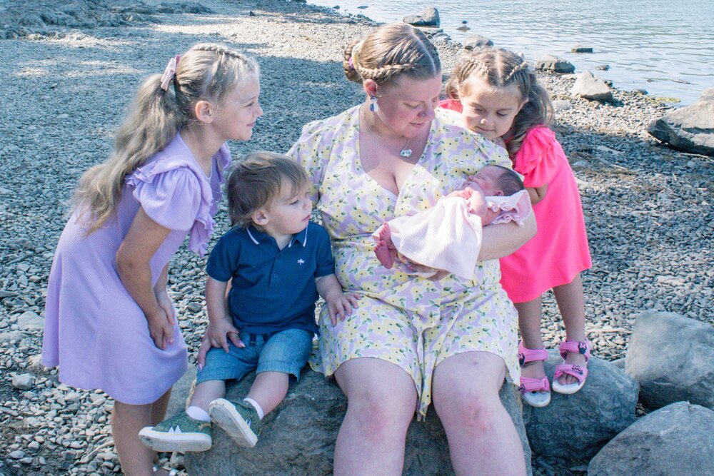 Naomi is holding baby Peanut and the older three children are around her. All are looking at the baby. They are on the lakeshore of Derwentwater.