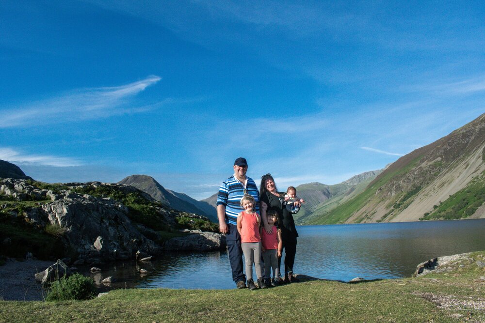 The Hassan family are on a grassy section of Wast Water lakeshore with Wast Water and the Lakeland fells in the background.