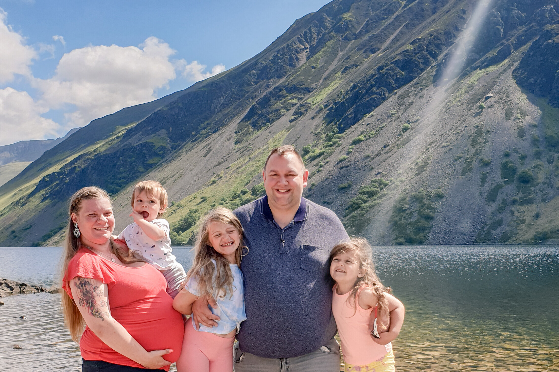 The Hassan family are on a grassy section of Wast Water lakeshore with Wast Water and the Lakeland fells in the background.