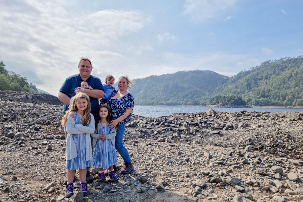 The Hassan family are stood on a rocky area of lakeshore with Thirlmere and Lakeland fells behind them.