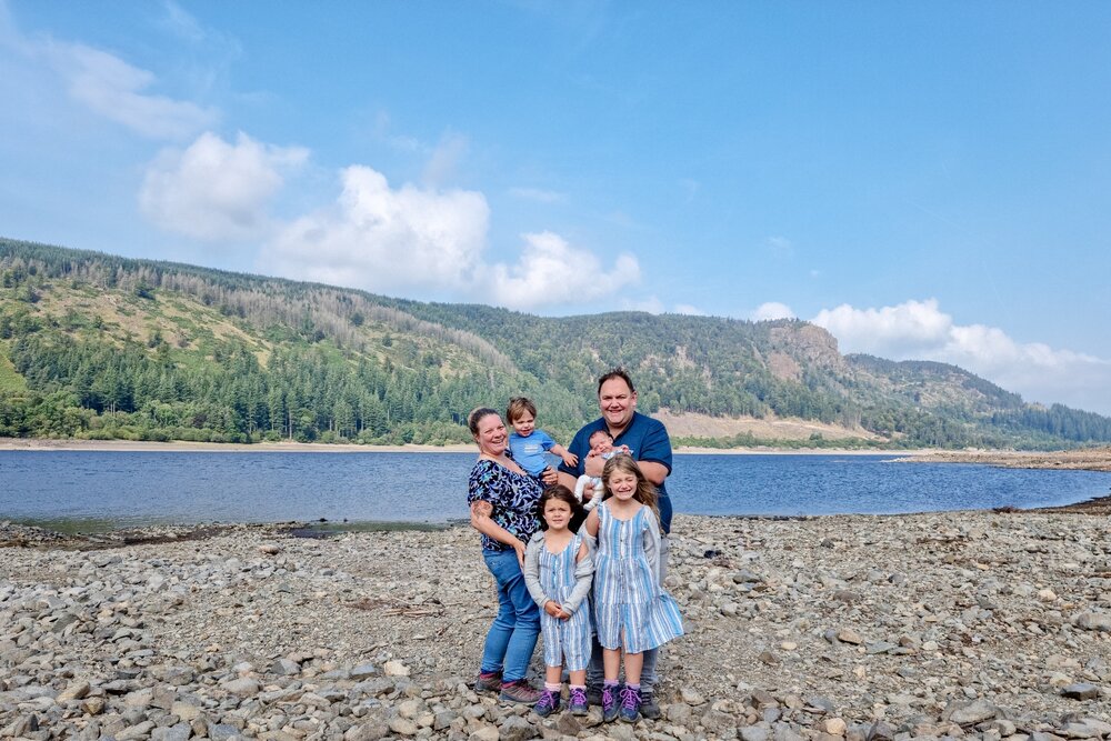 The Hassan family are stood on a rocky area of lakeshore with Thirlmere and Lakeland fells behind them.
