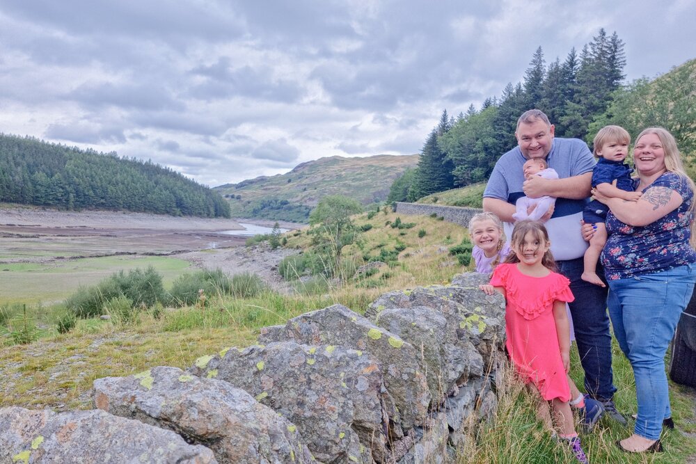 The Hassan family are together in the right of the image, stood next to an old boundary wall with Haweswater and lakeland fells in the distance behind them to the left of the image.