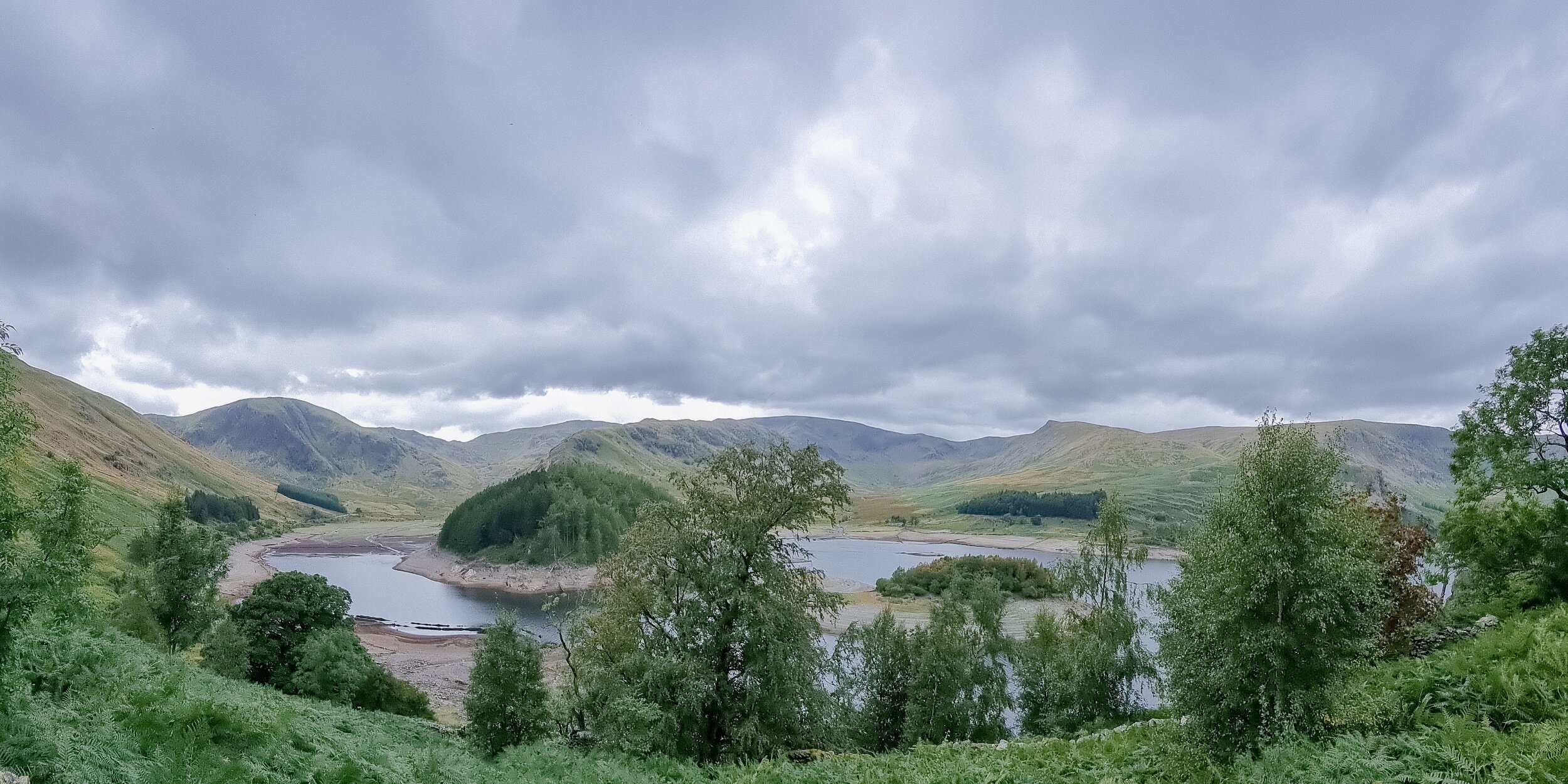 A panoramic image of Haweswater. You can see an island in the centre and some lakeland fells in the background. The lake is very low.