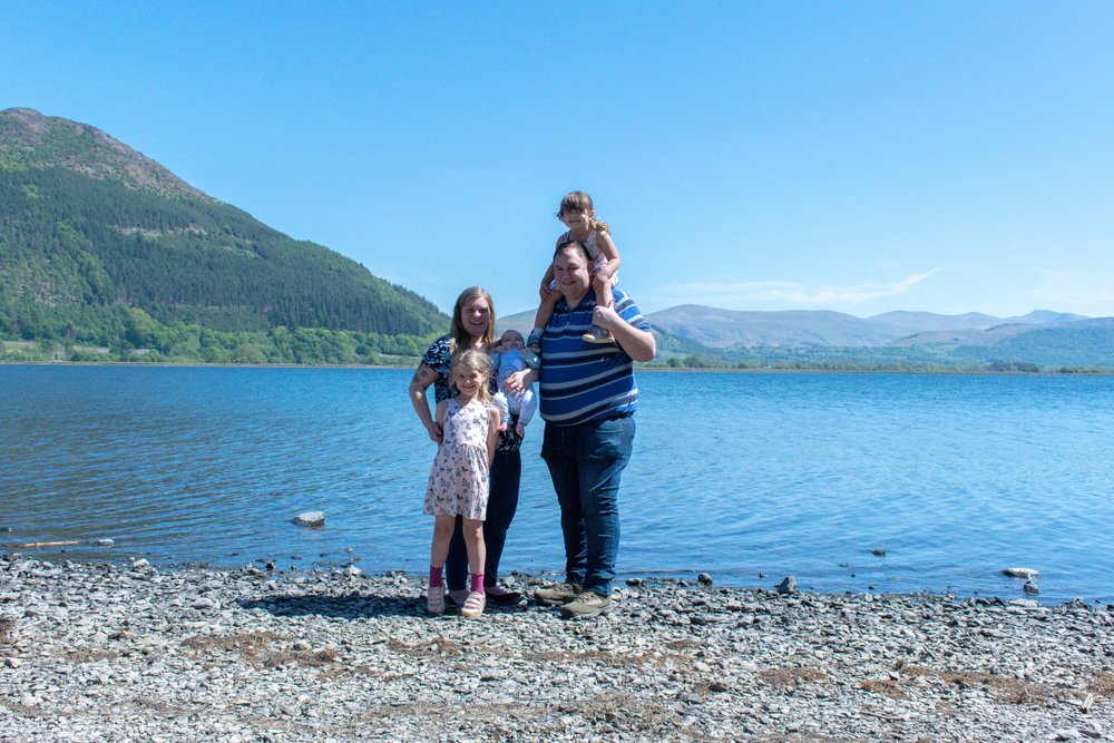 The Hassan family are stood together on the lakeshore of Bassenthwaite with the fells in the background to celebrate Munchkin's birth.