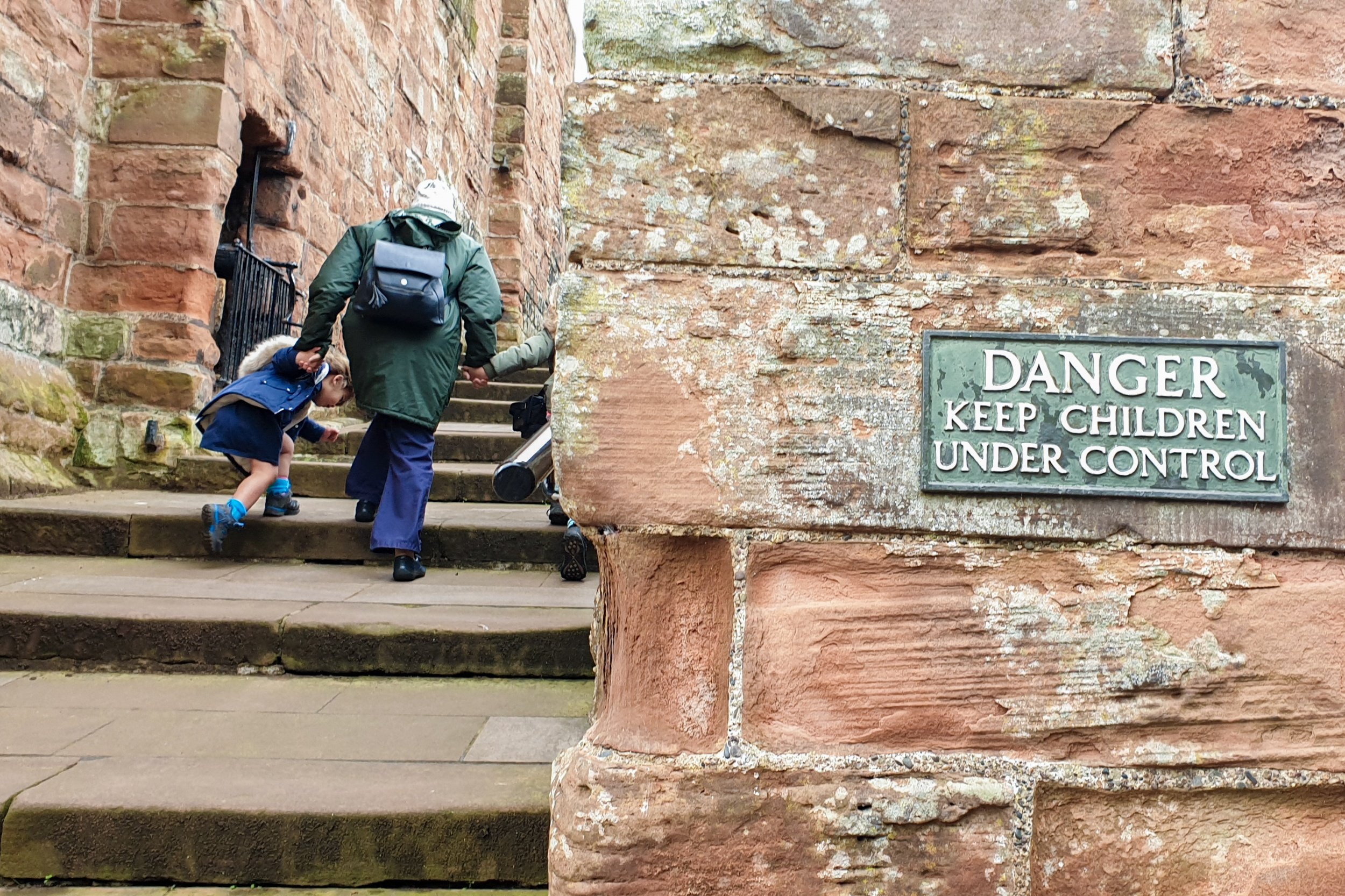 Squidgy and Pickle are holding their Grandma's hand and walking up the steps inside Carlisle castle. A sign on the wall beside them says, "Danger, keep children under control".