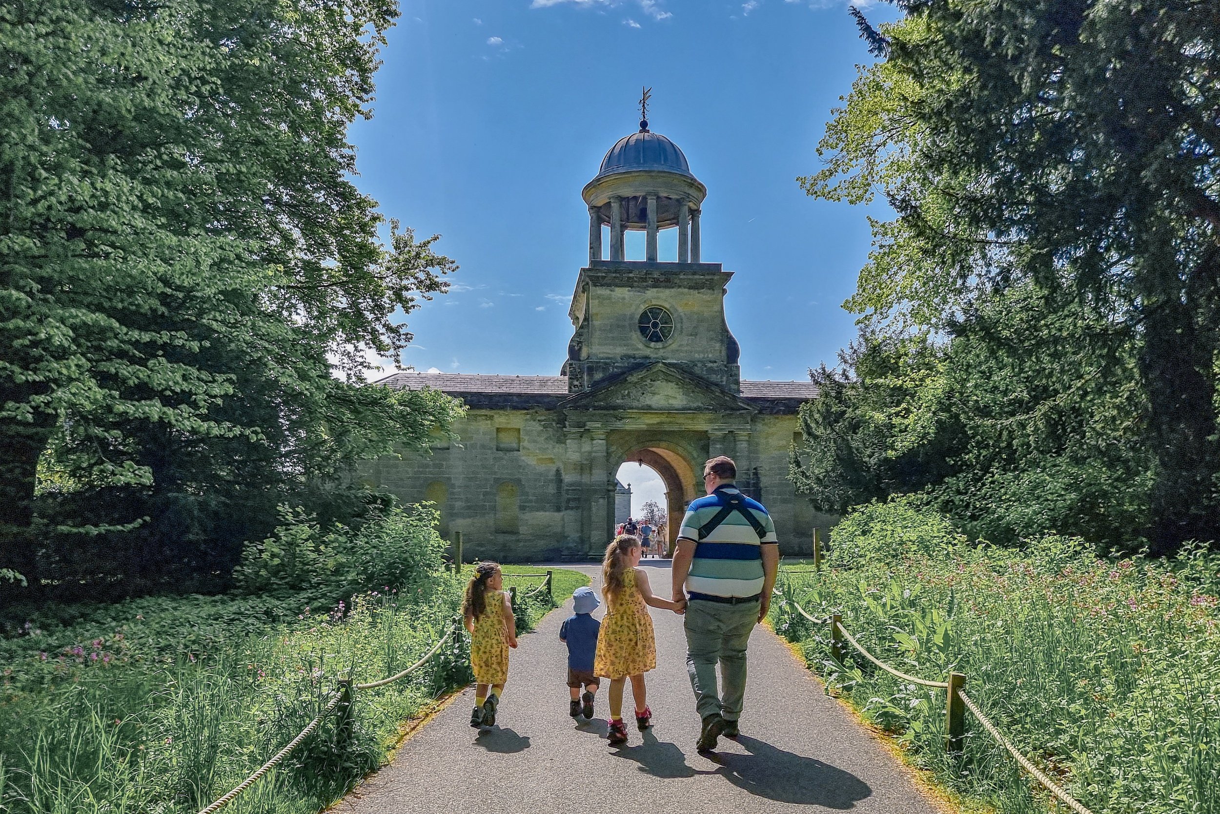 John is baby wearing, while holding Squidgy's hand and walking alongside Pickle and Munchkin. they are on a path with the Wallington clocktower ahead of them.