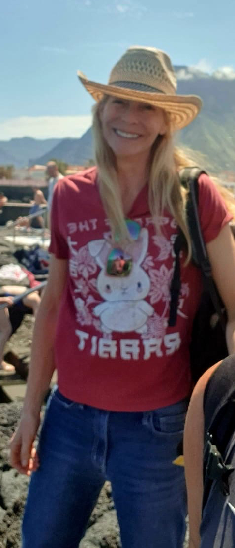 photo of tall blonde artist with Gran Canaria volcanoes and mountains in the background. The woman is wearing a straw hat and a red shirt and blue jeans. She is smiling widely. The sky is blue and it is a sunny day.