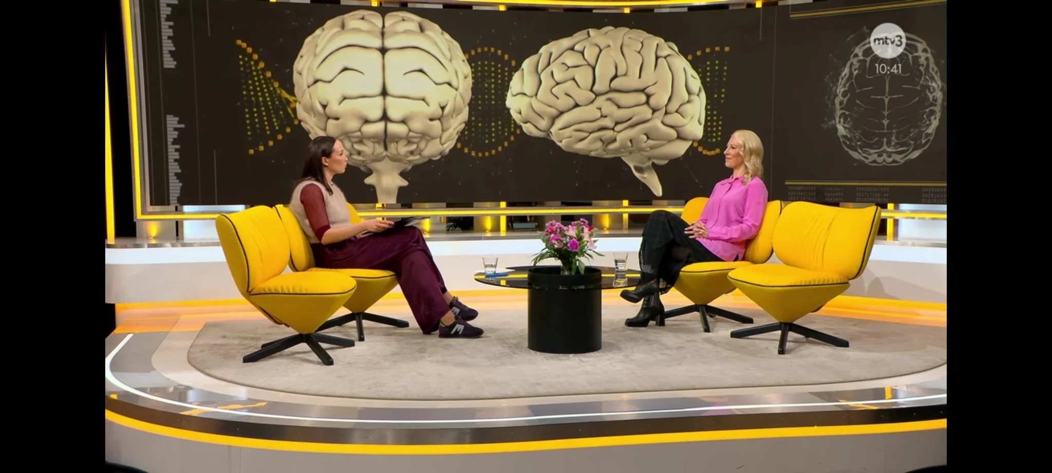 Kaksi naista keskustelee studiossa, taustalla suuri näyttökuva aivoista.