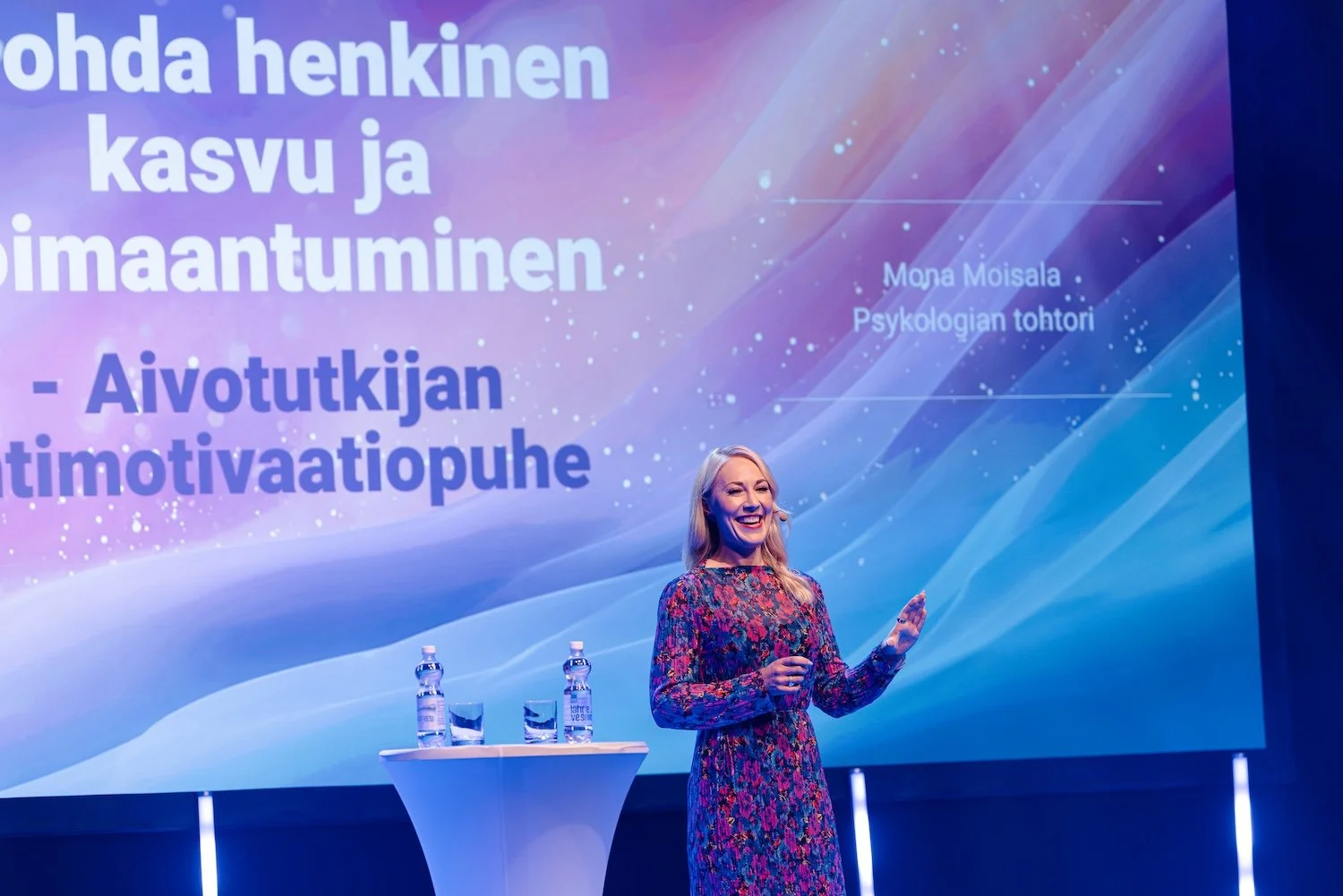 Mona puhumassa lavalla, taustalla suuri diaesitys.
