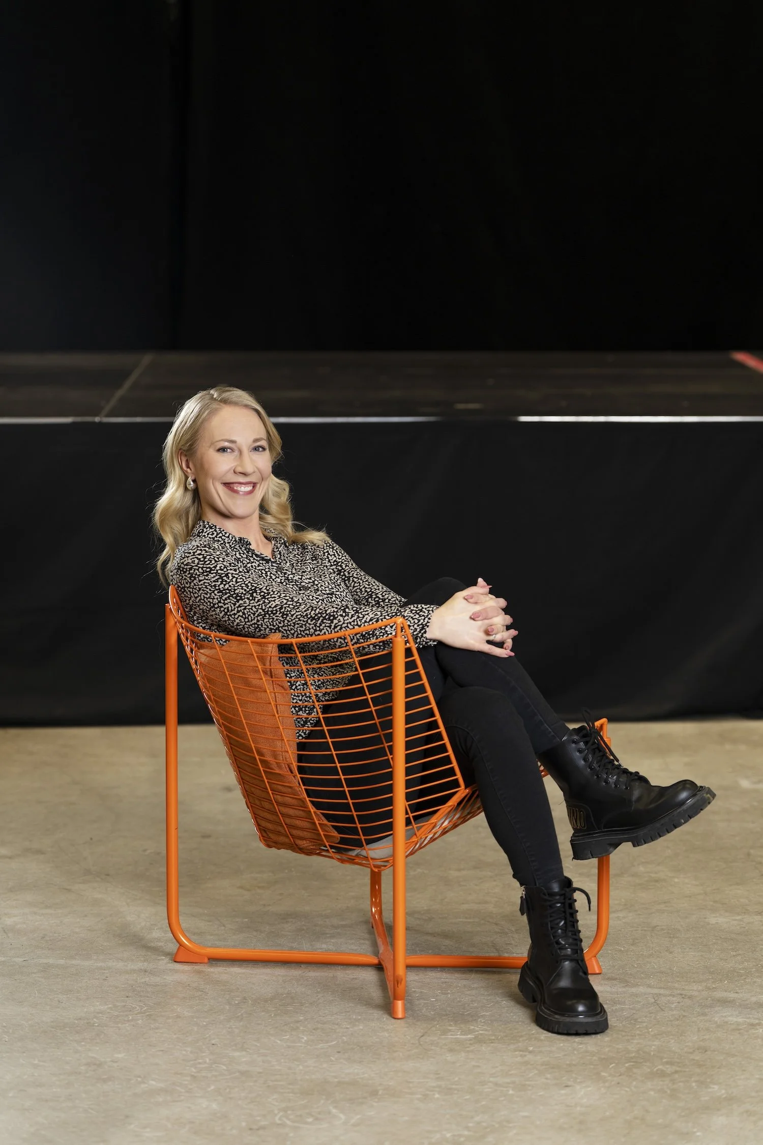 Mona istuu oranssissa metallituolissa ja hymyilee kameralle, musta tausta ja lattia puuta.