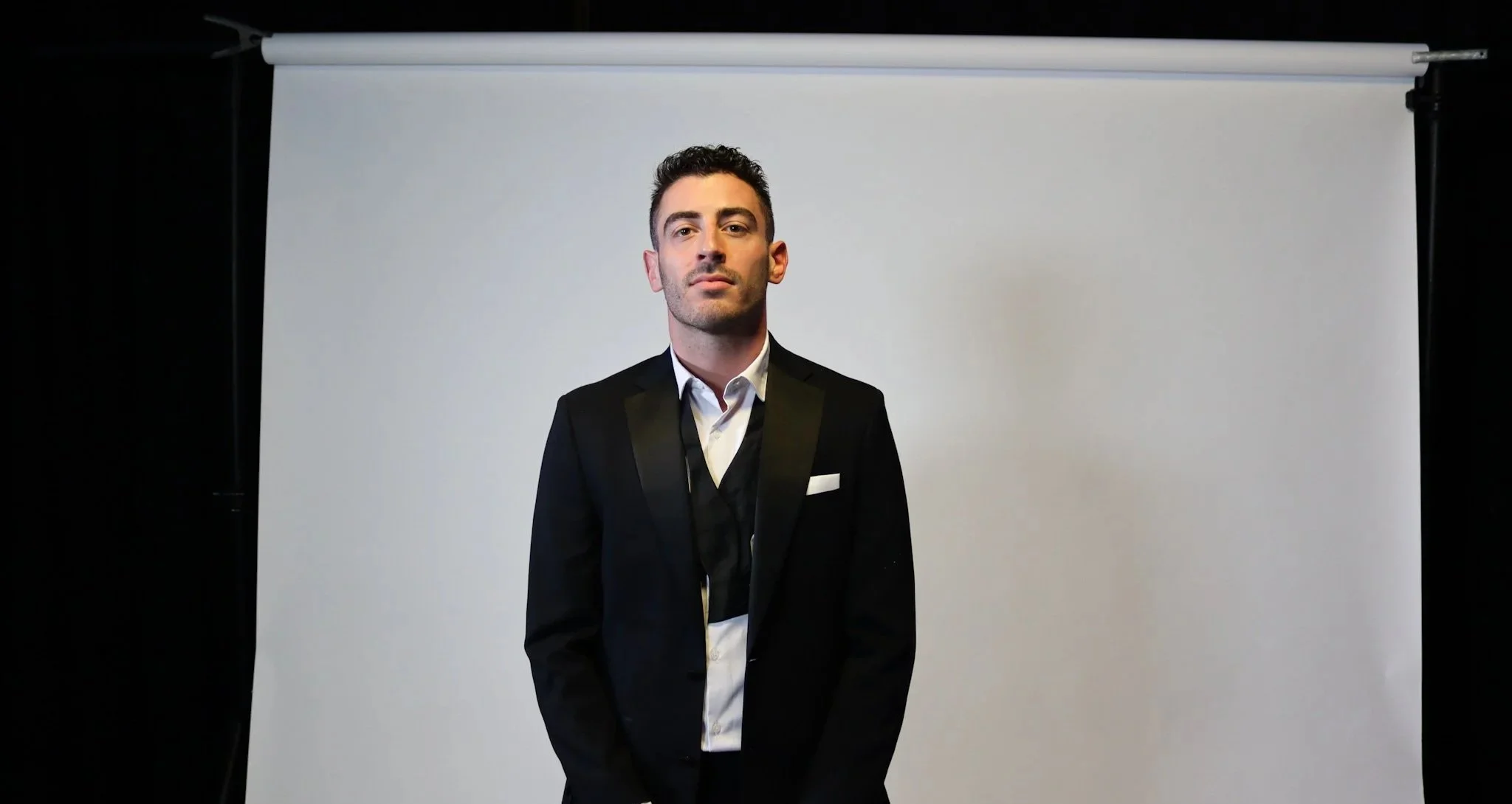 A man in a black tuxedo with a white shirt stands in front of a plain white backdrop.