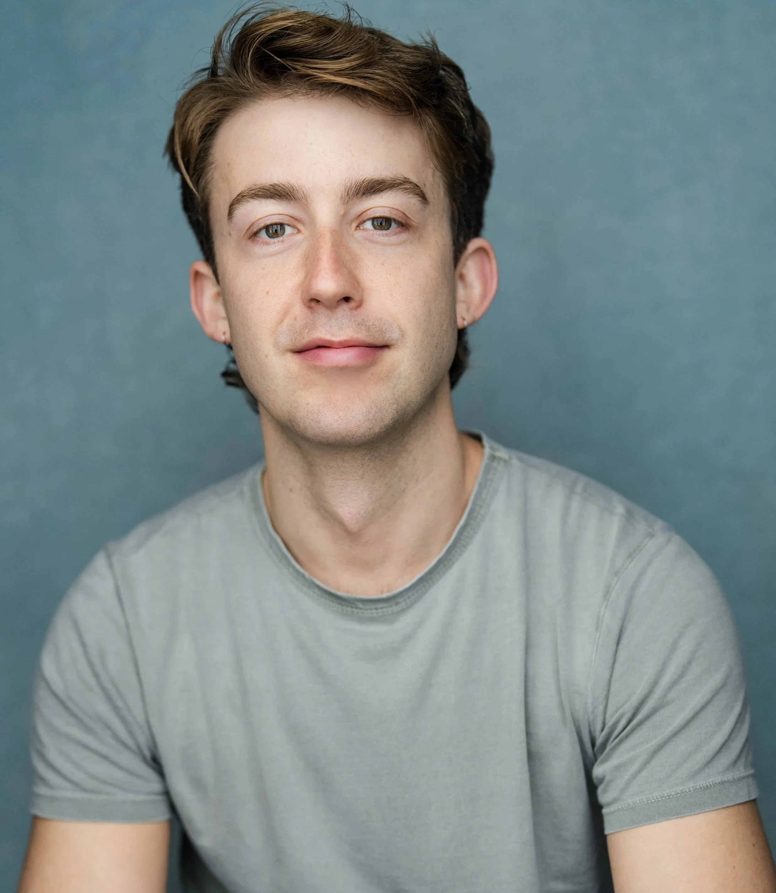 Portrait of a young man with light skin, brown hair, and blue eyes, wearing a gray t-shirt, against a blue background.