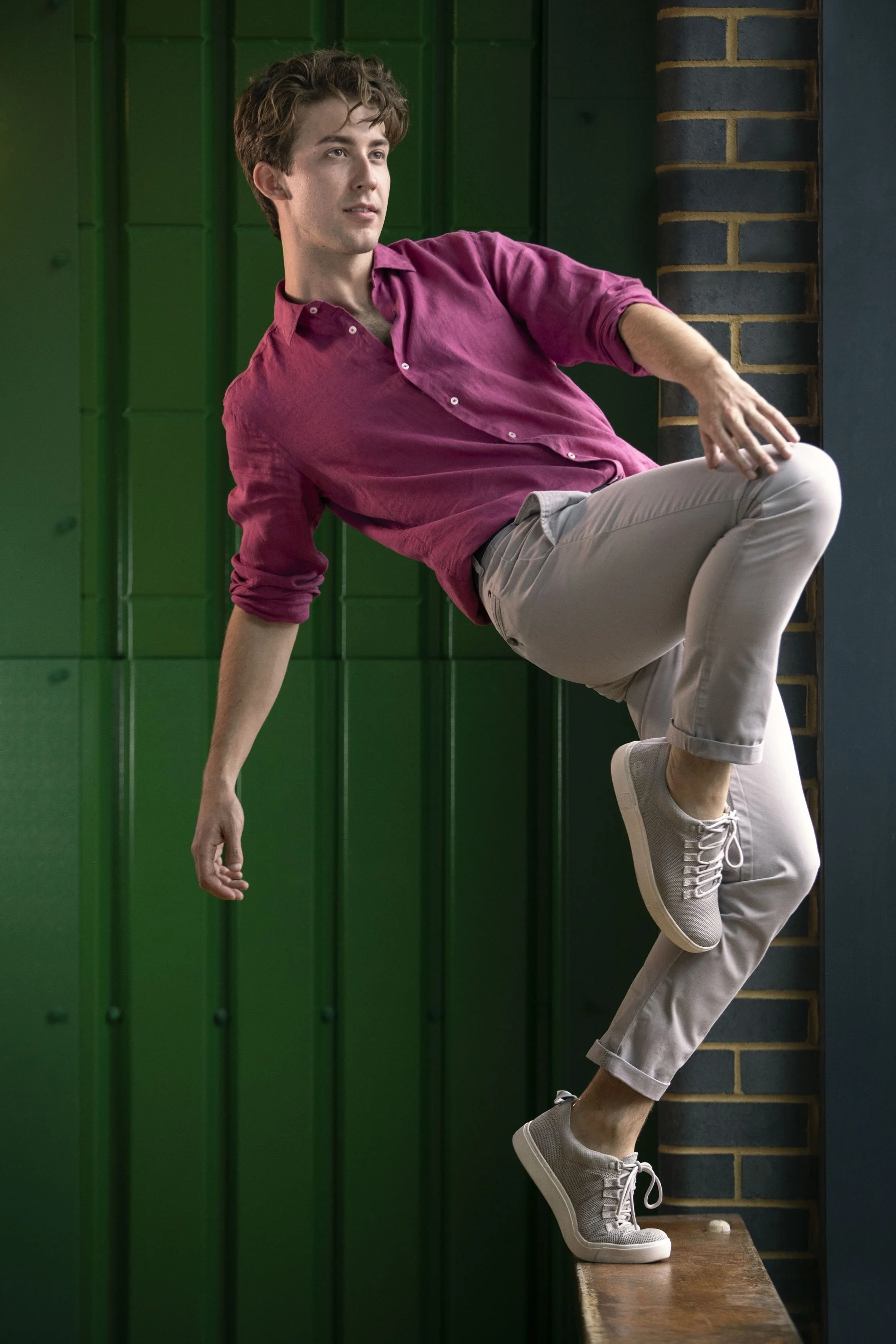 A young man in a pink button-up shirt and beige pants balancing on a wooden ledge with one foot, against a background of green panels and brick wall.