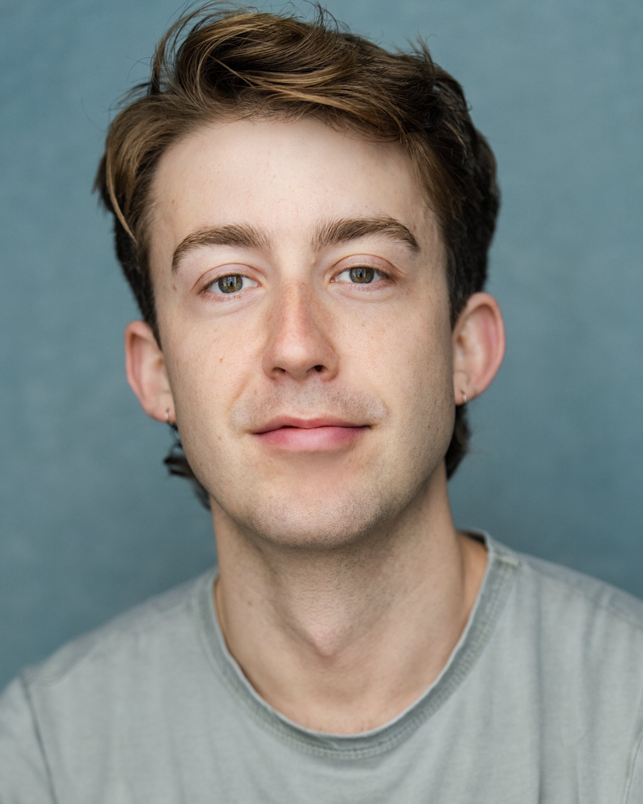Headshot of a man with short brown hair, light eyes and a calm expression, wearing a light grey T-shirt in front of a soft blue backdrop.
