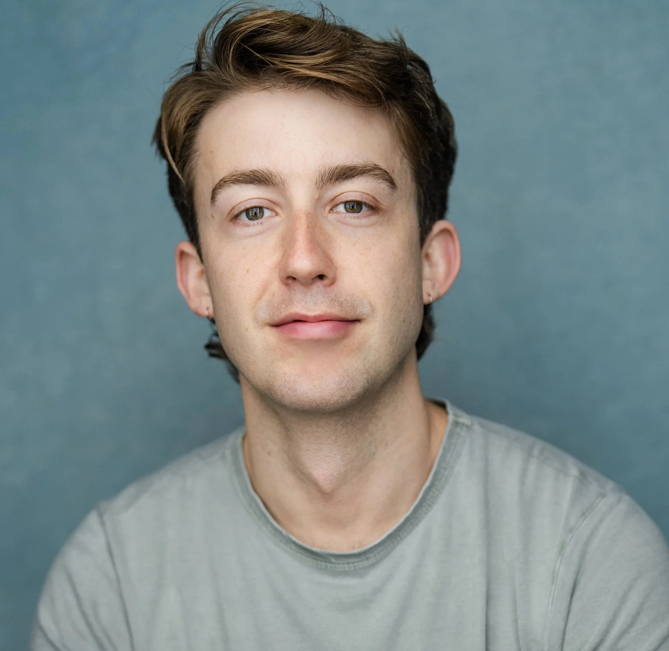 Portrait of a young man with brown hair and blue eyes, wearing a light gray T-shirt, against a blue background.