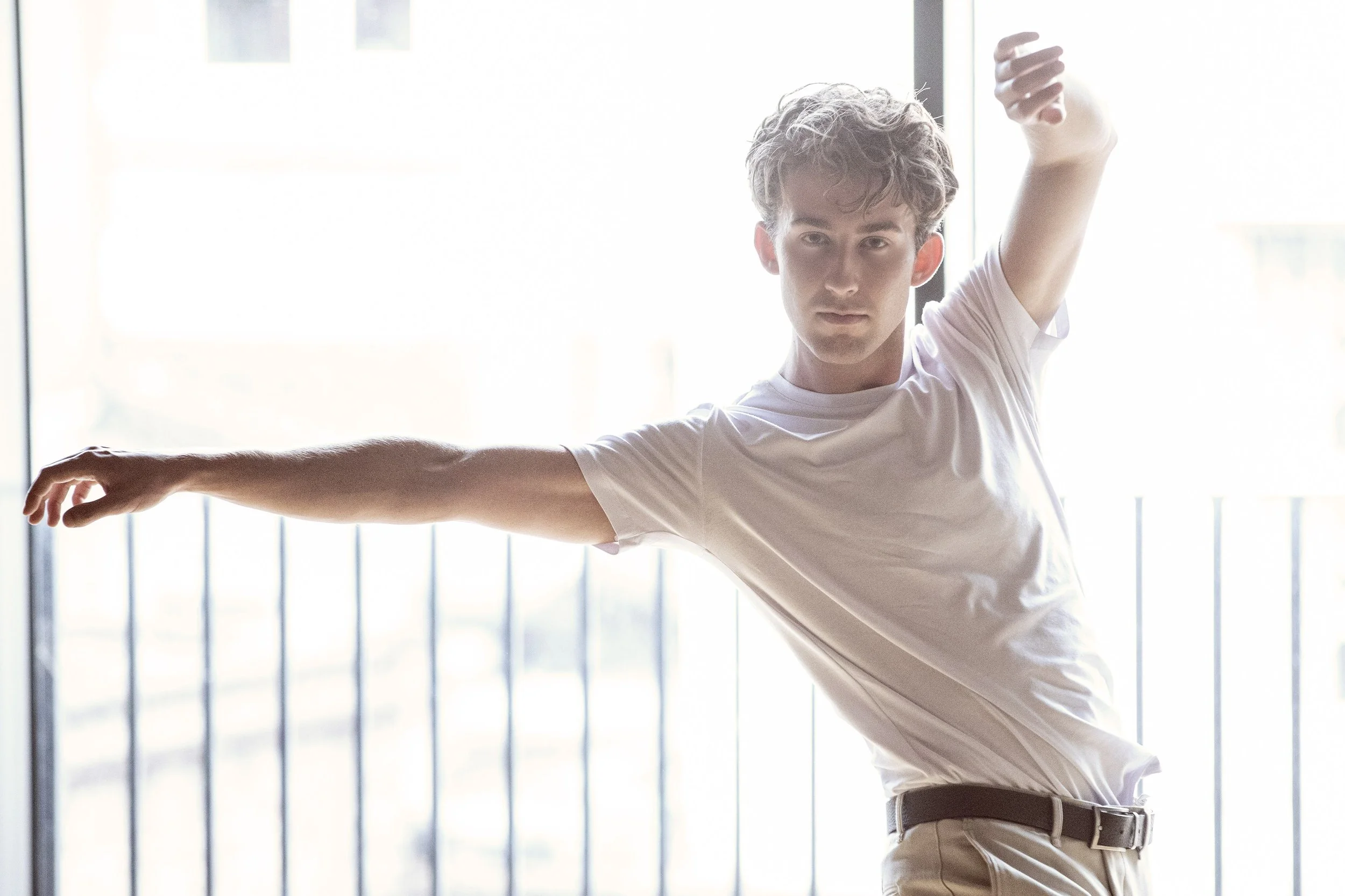 A young man with curly hair stretching with arms extended, standing indoors near large windows with blinds.