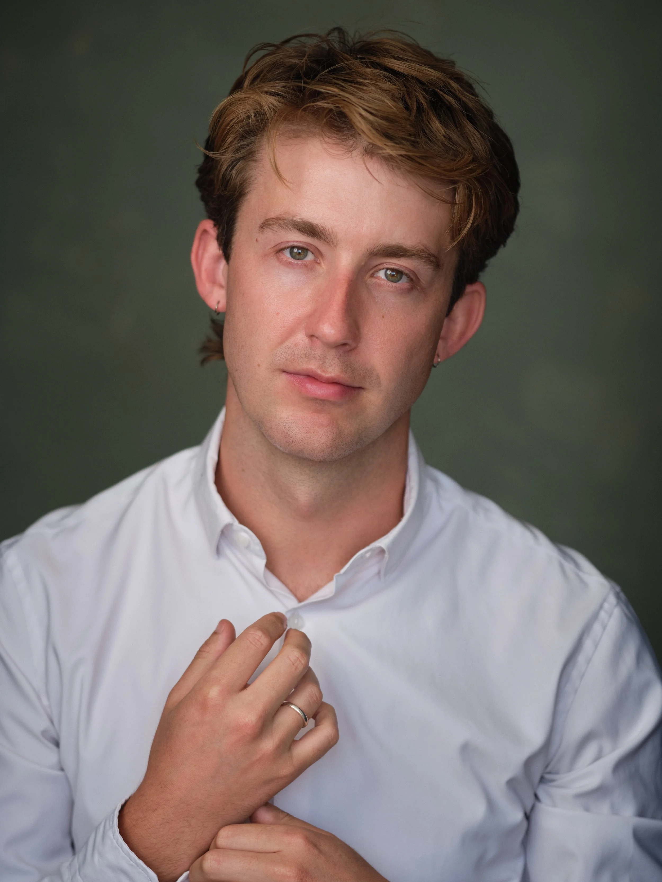 A young man with light skin and brown hair adjusting his white dress shirt.