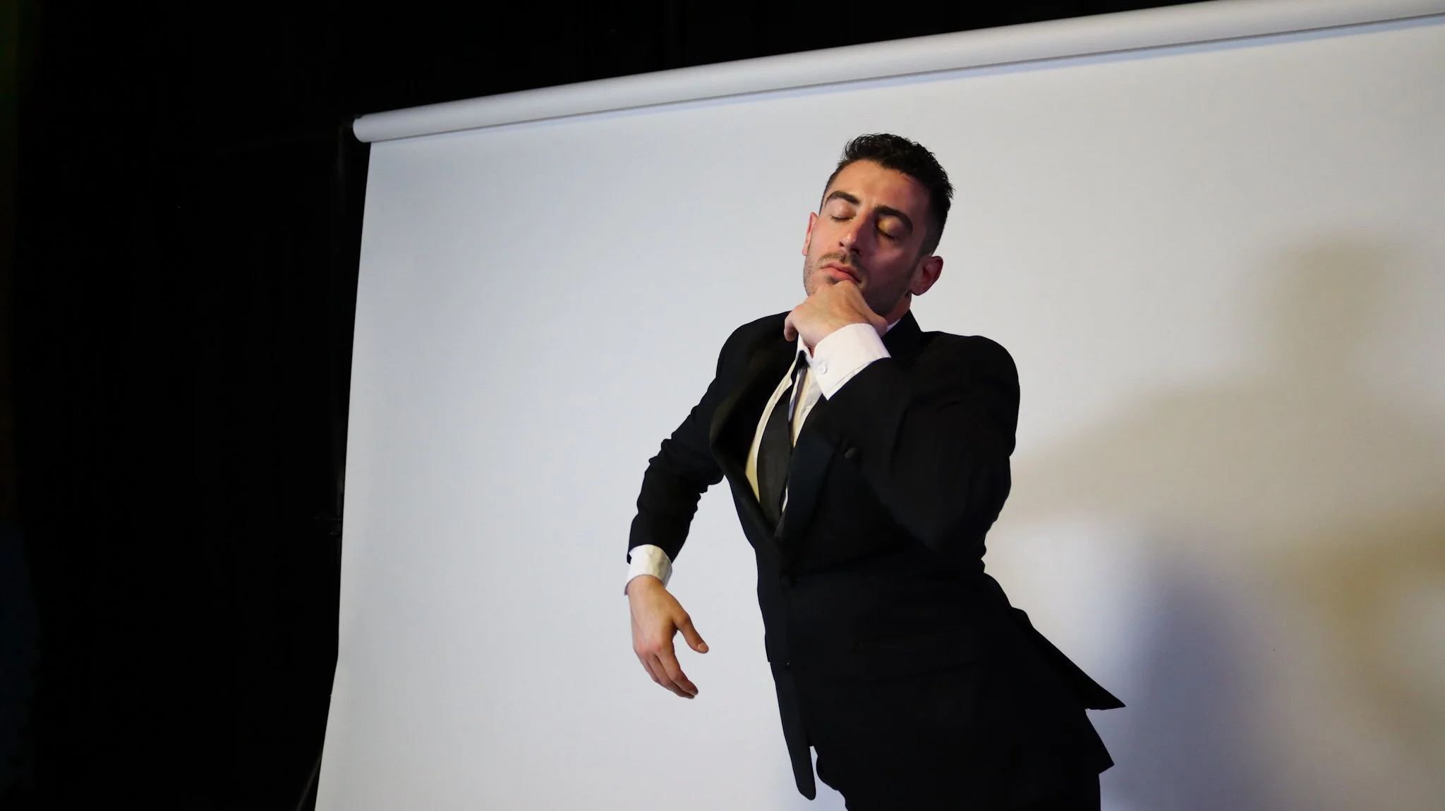 A man in a black suit and white shirt posing with his hand on his chin in front of a white backdrop.