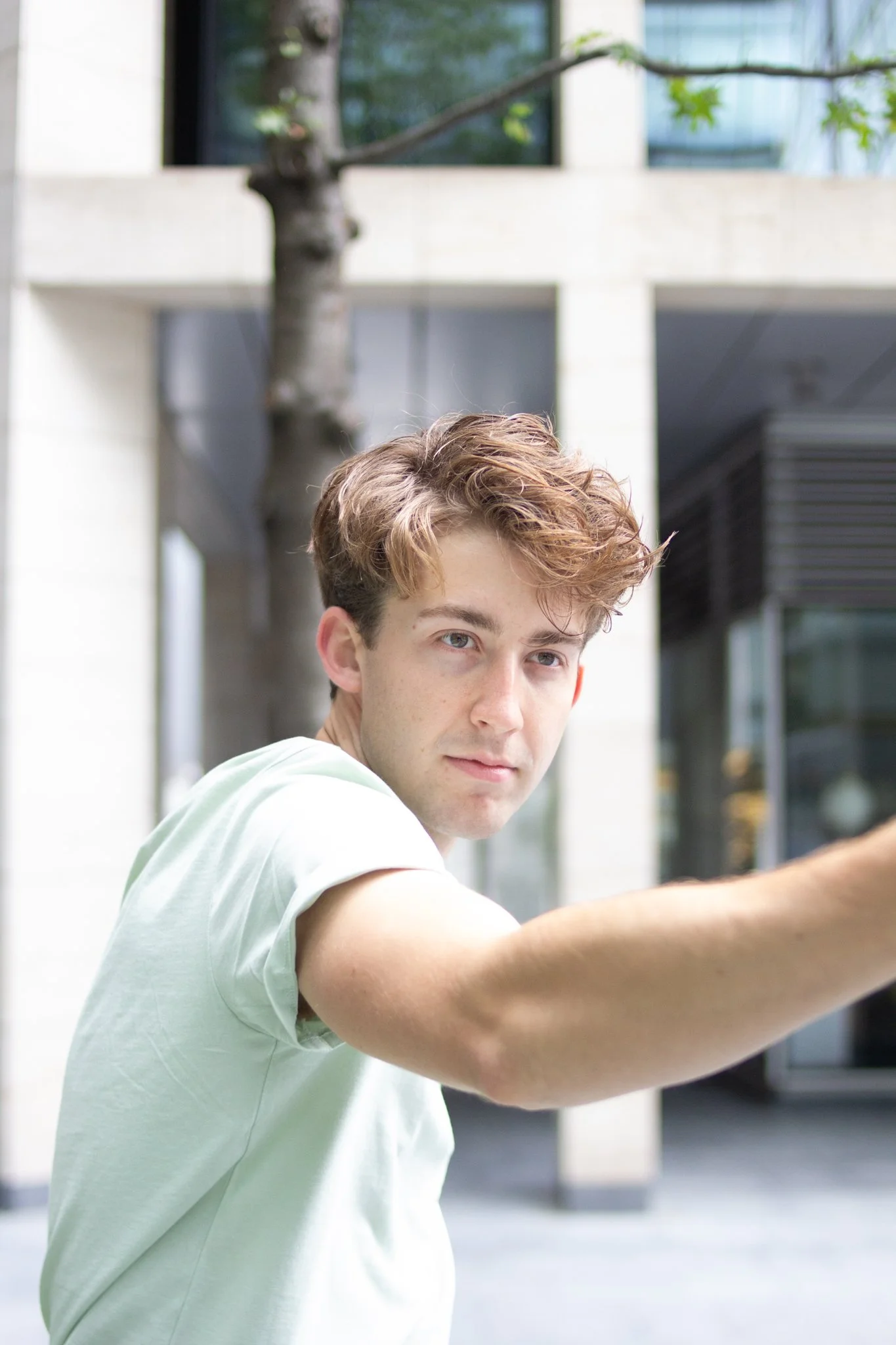 A young man with curly brown hair and light skin reaches out with his arm in front of him outdoors, with modern building architecture and a tree in the background.