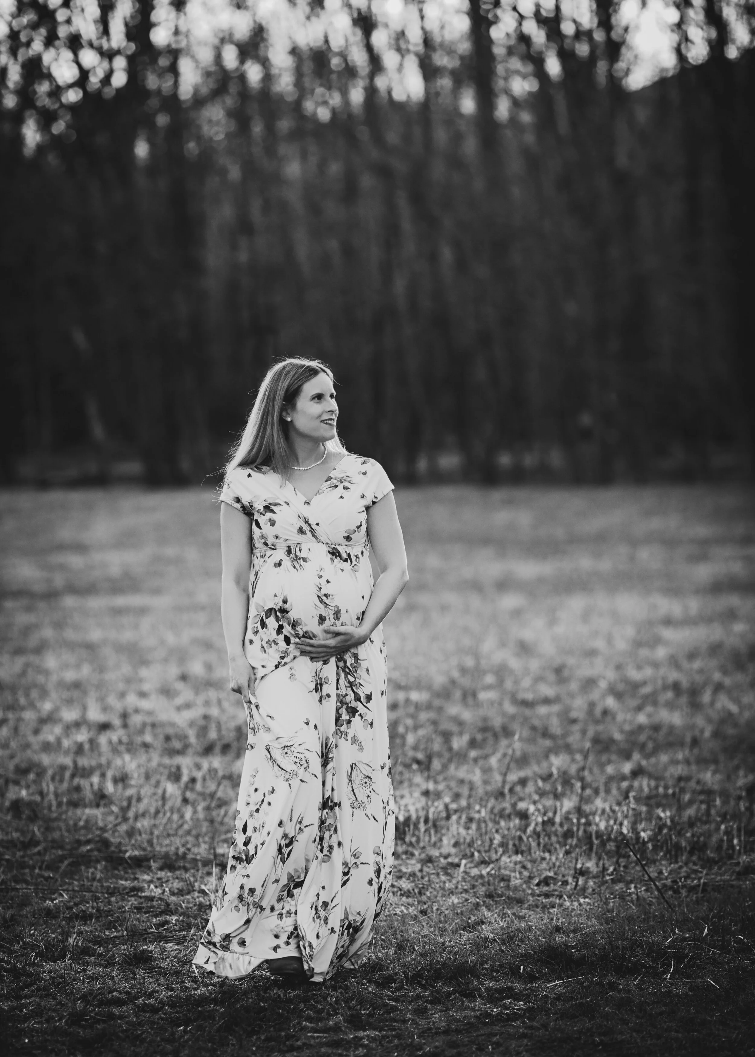Pregnant woman wearing a floral dress holding her belly, standing in a field with trees in the background, black and white photo.