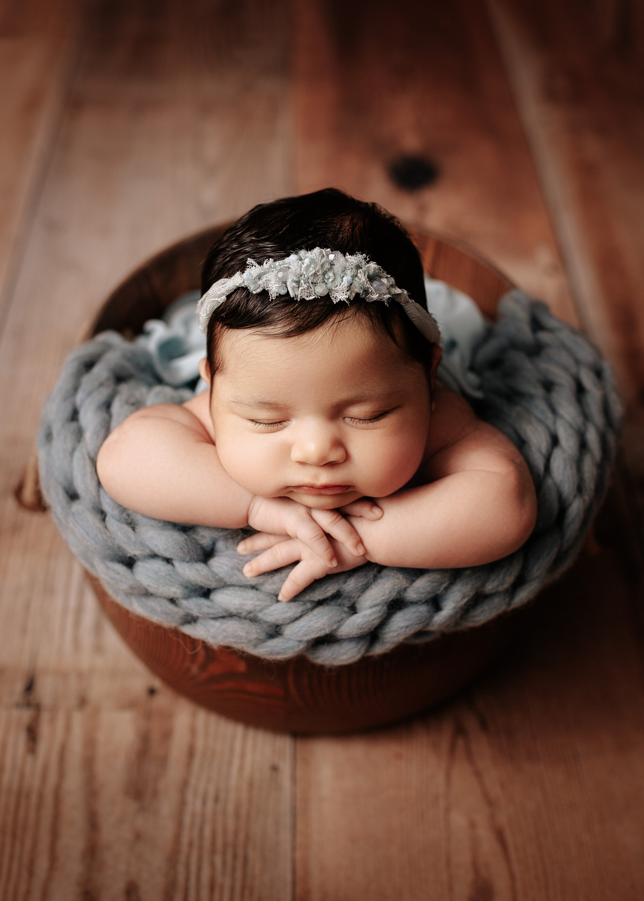 Newborn baby sleeping in a wooden bowl, wrapped in a blue knitted blanket, wearing a floral headband on a wooden floor.