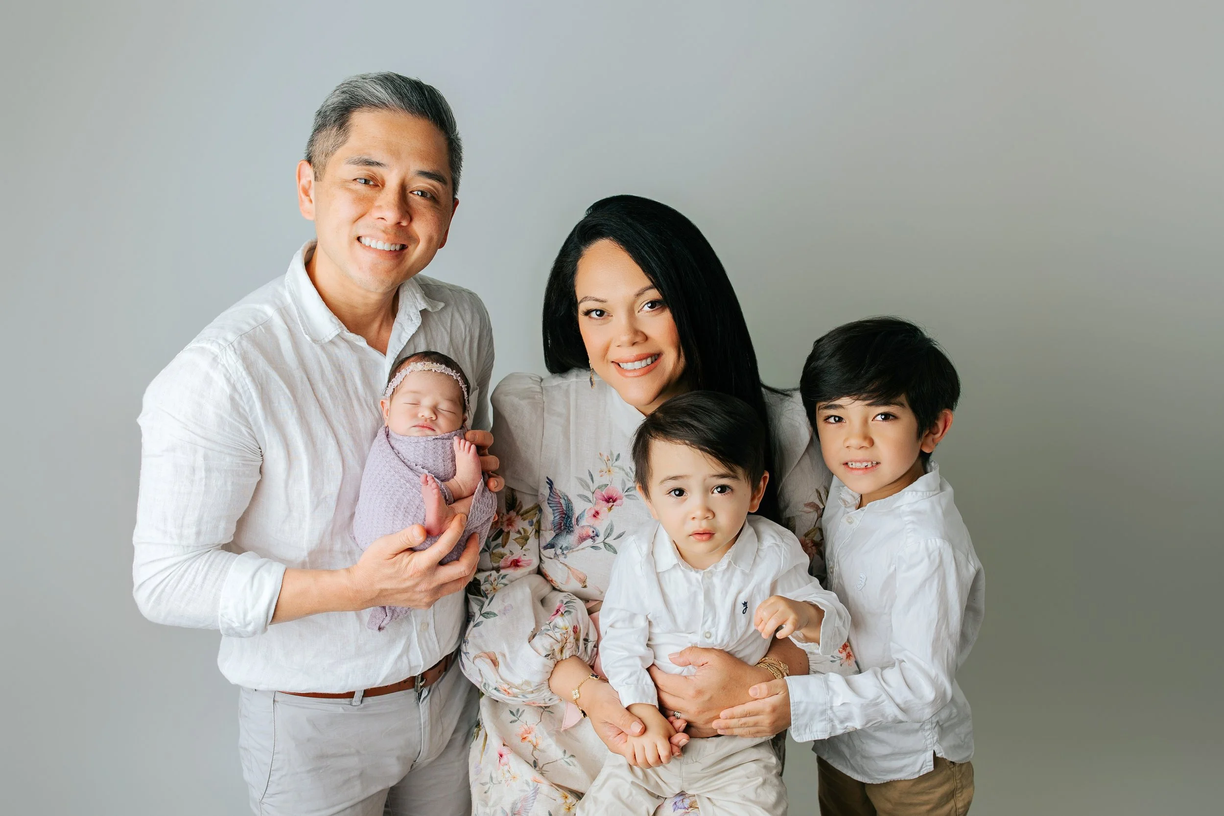 A family of six, including a man, woman, three children, and a newborn baby, posing together in front of a gray background, smiling and dressed in light-colored and floral clothing.