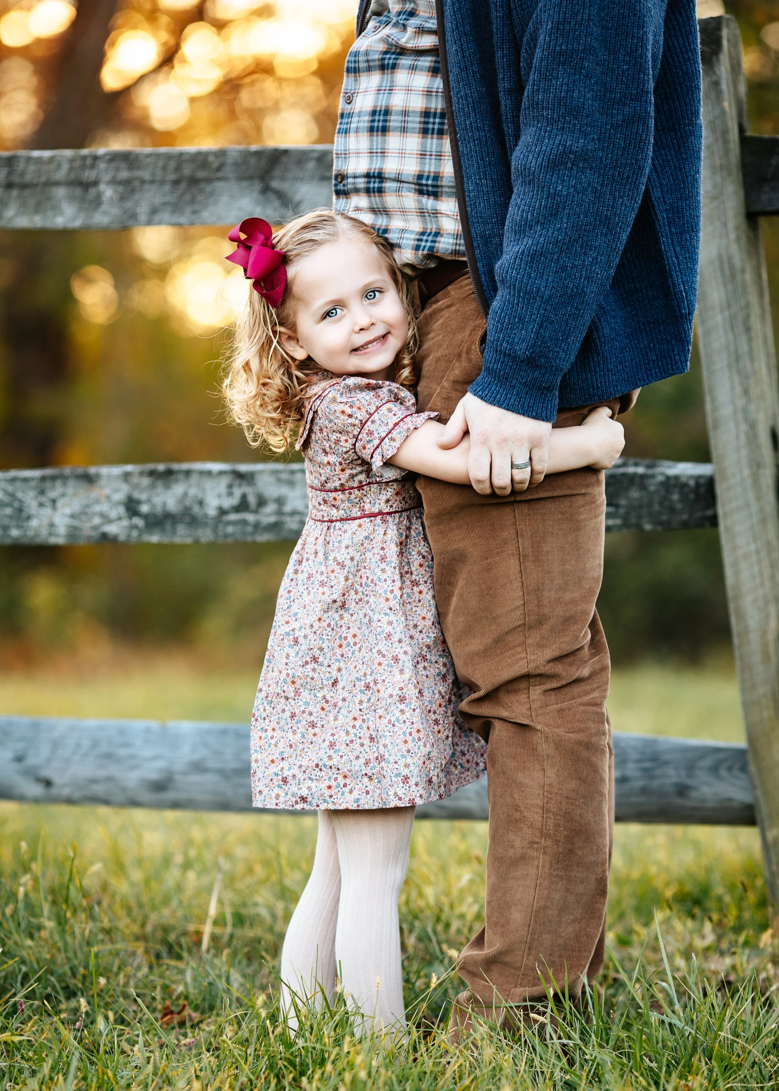 Blonde girl with red bow hugging dad's legs in Morrstown NJ looking at the NJ photographer camera